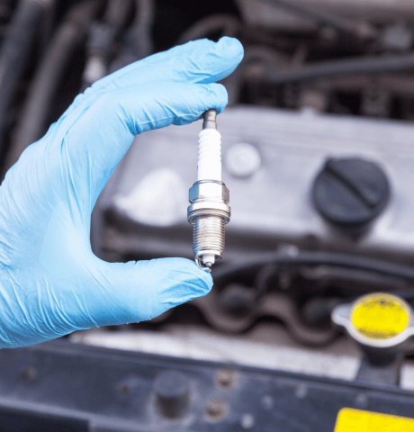 Technician holding a spark plug above an open vehicle engine bay.