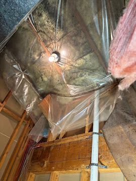 Attic with exposed beams, foil insulation, and a hanging light bulb above a white pipe.