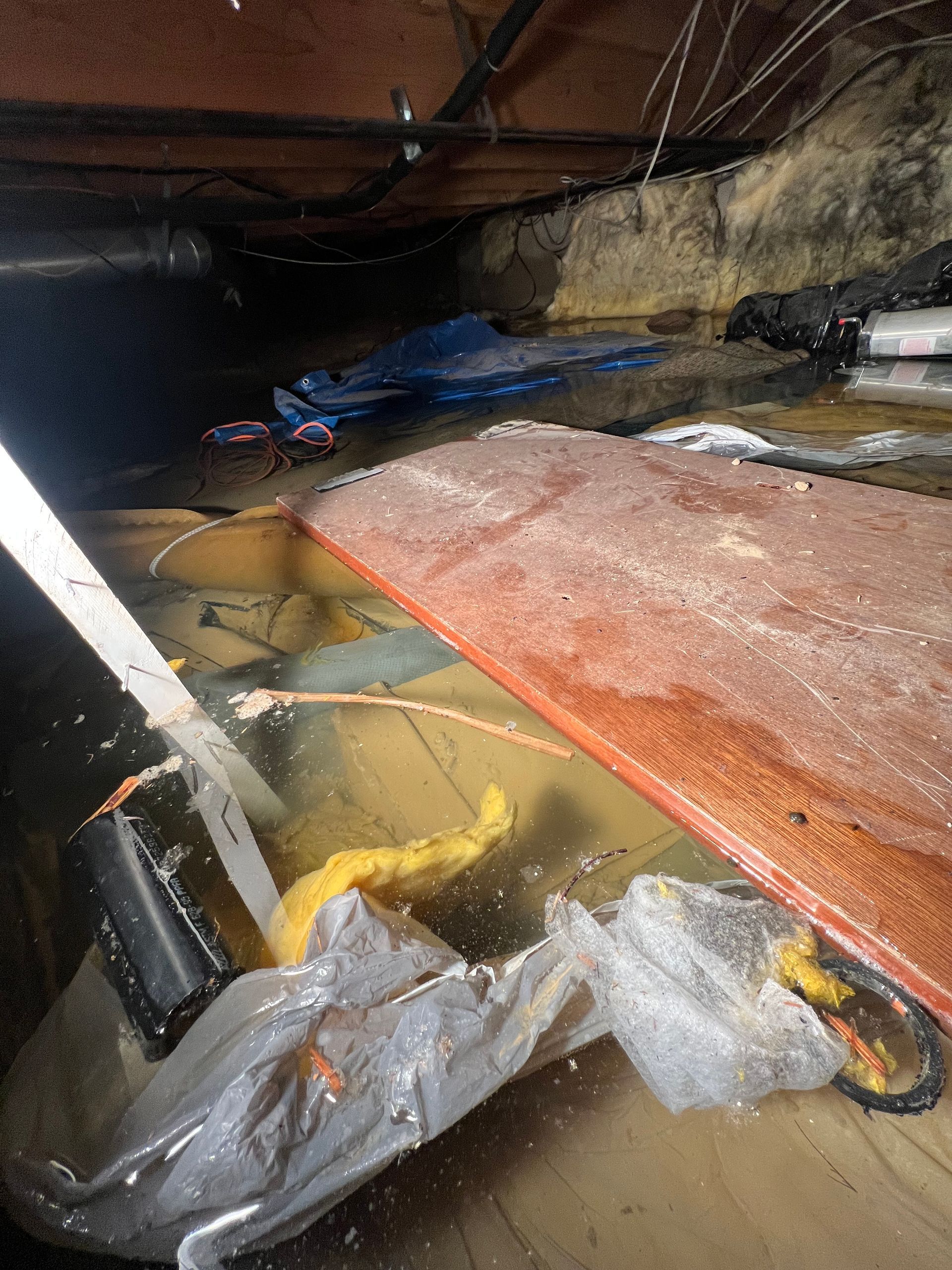 Flooded crawlspace with standing water, insulation, pipes, and debris under a wooden floor
