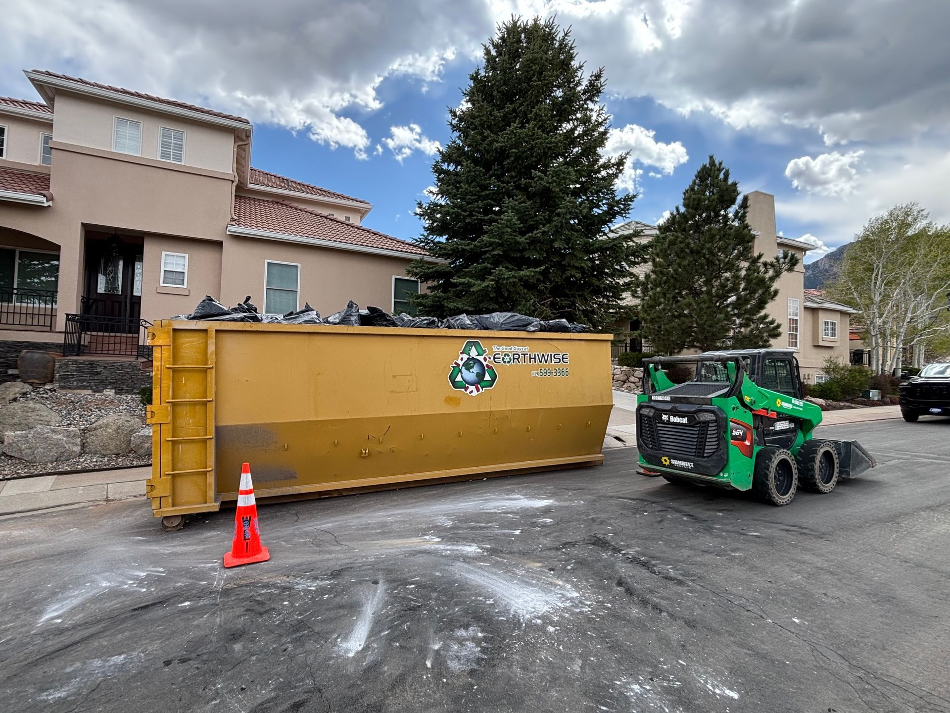 Yellow dumpster on wet pavement beside a green skid steer in a suburban parking lot