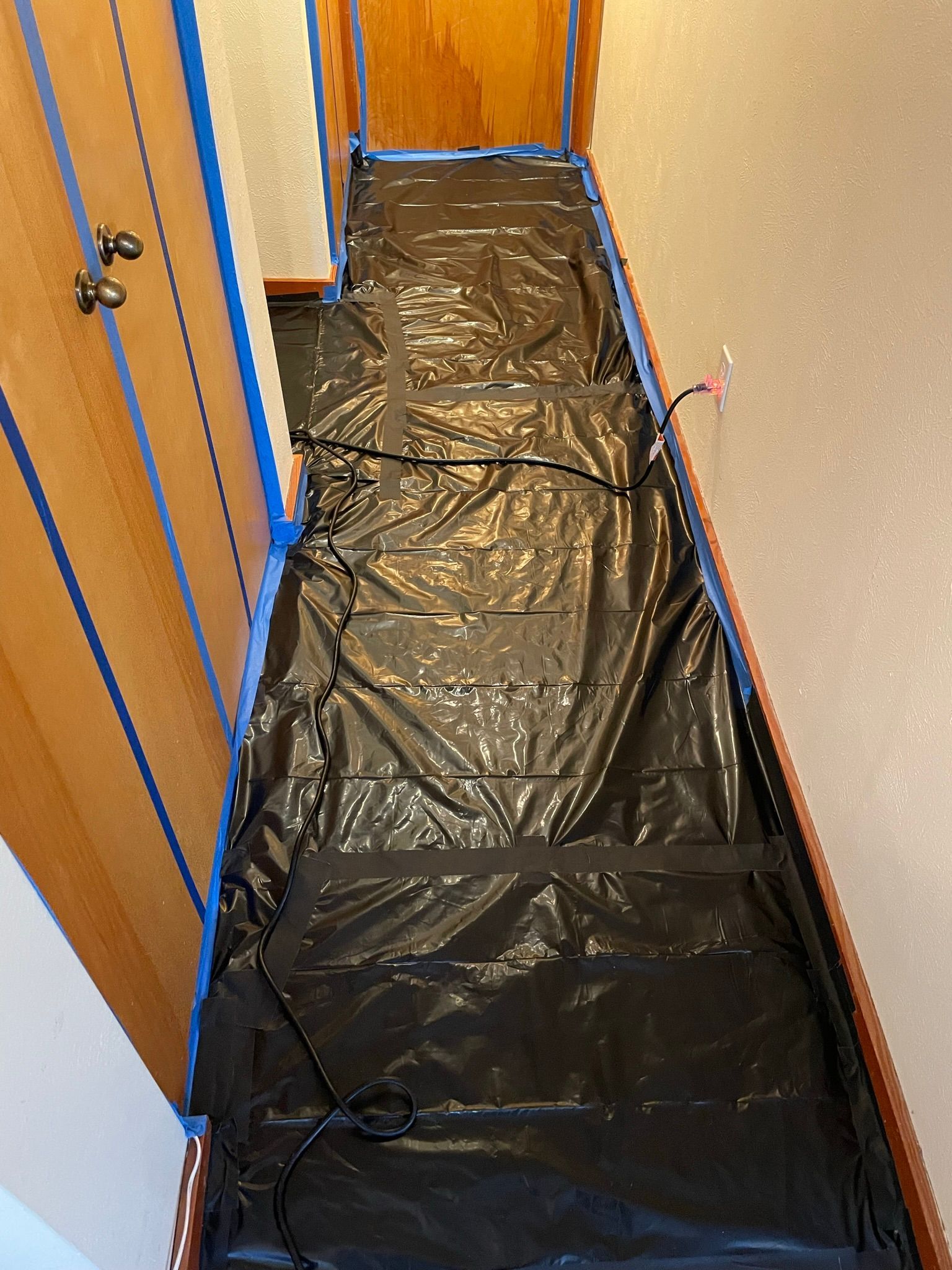 Hallway floor covered with black plastic sheeting beside wooden cabinets and a beige wall