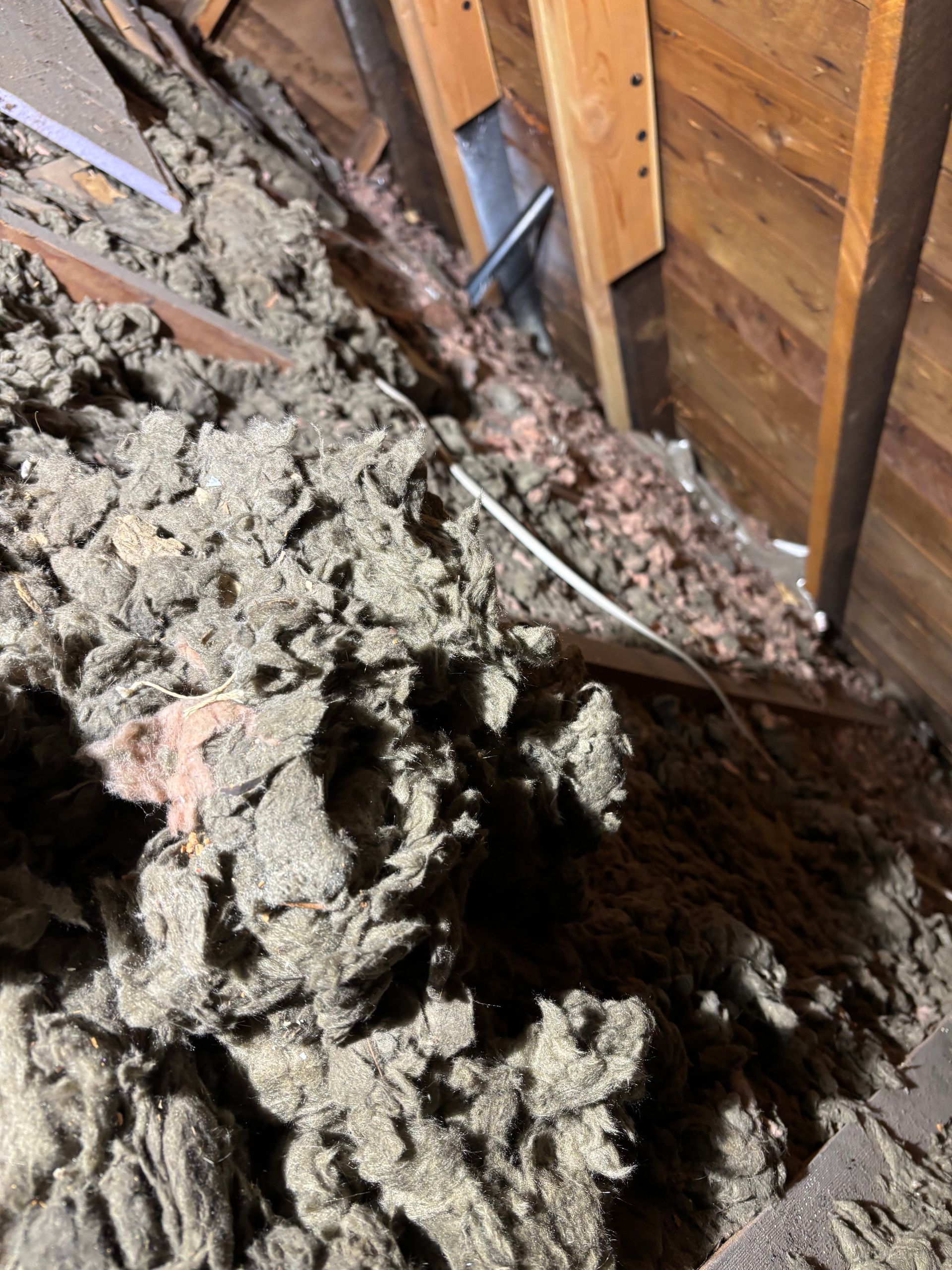 Insulation and debris piled in an attic beside exposed wooden rafters and wiring