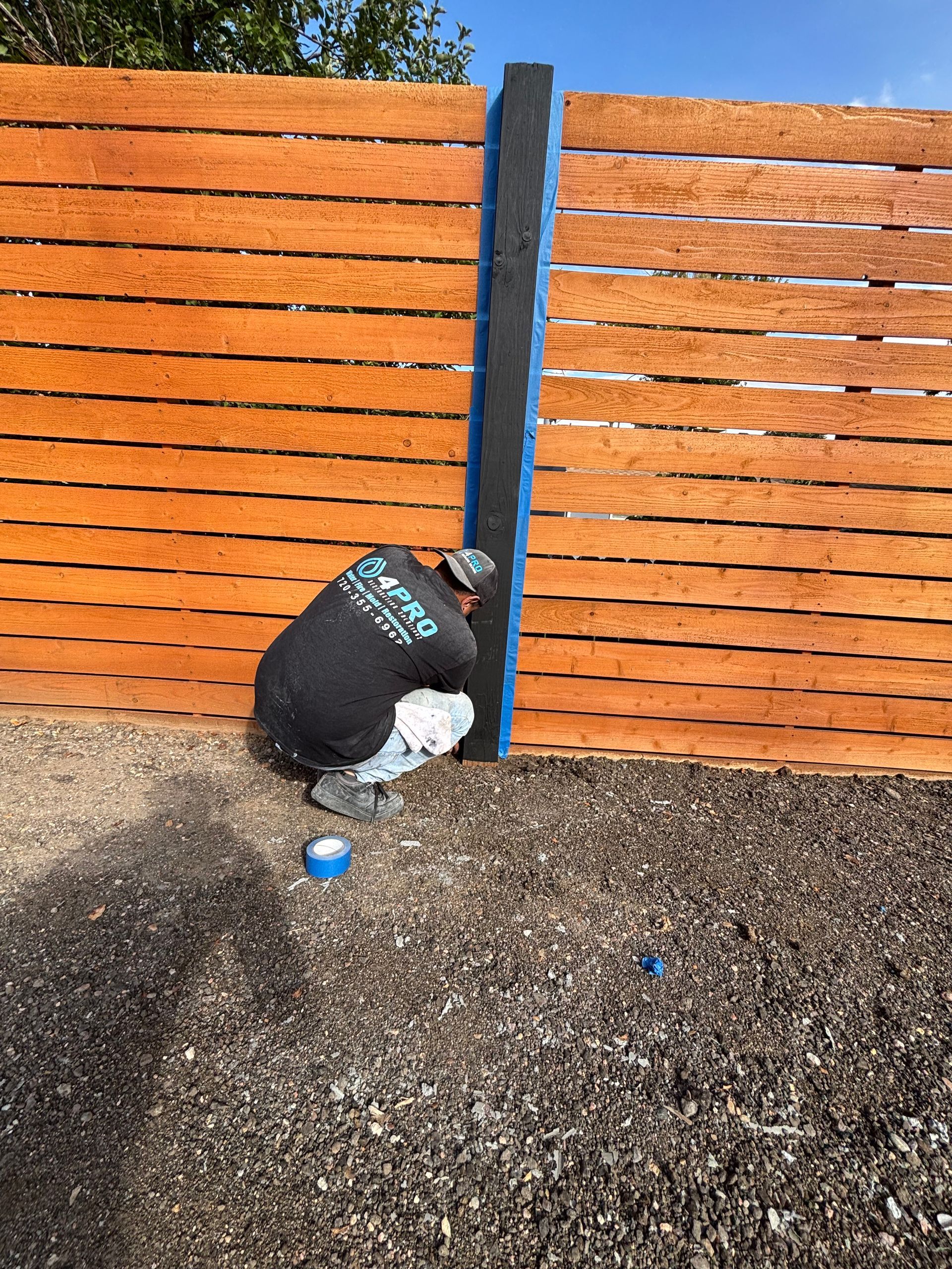Black trash bag leaning against a blue post in front of an orange wooden fence, with gravel below.