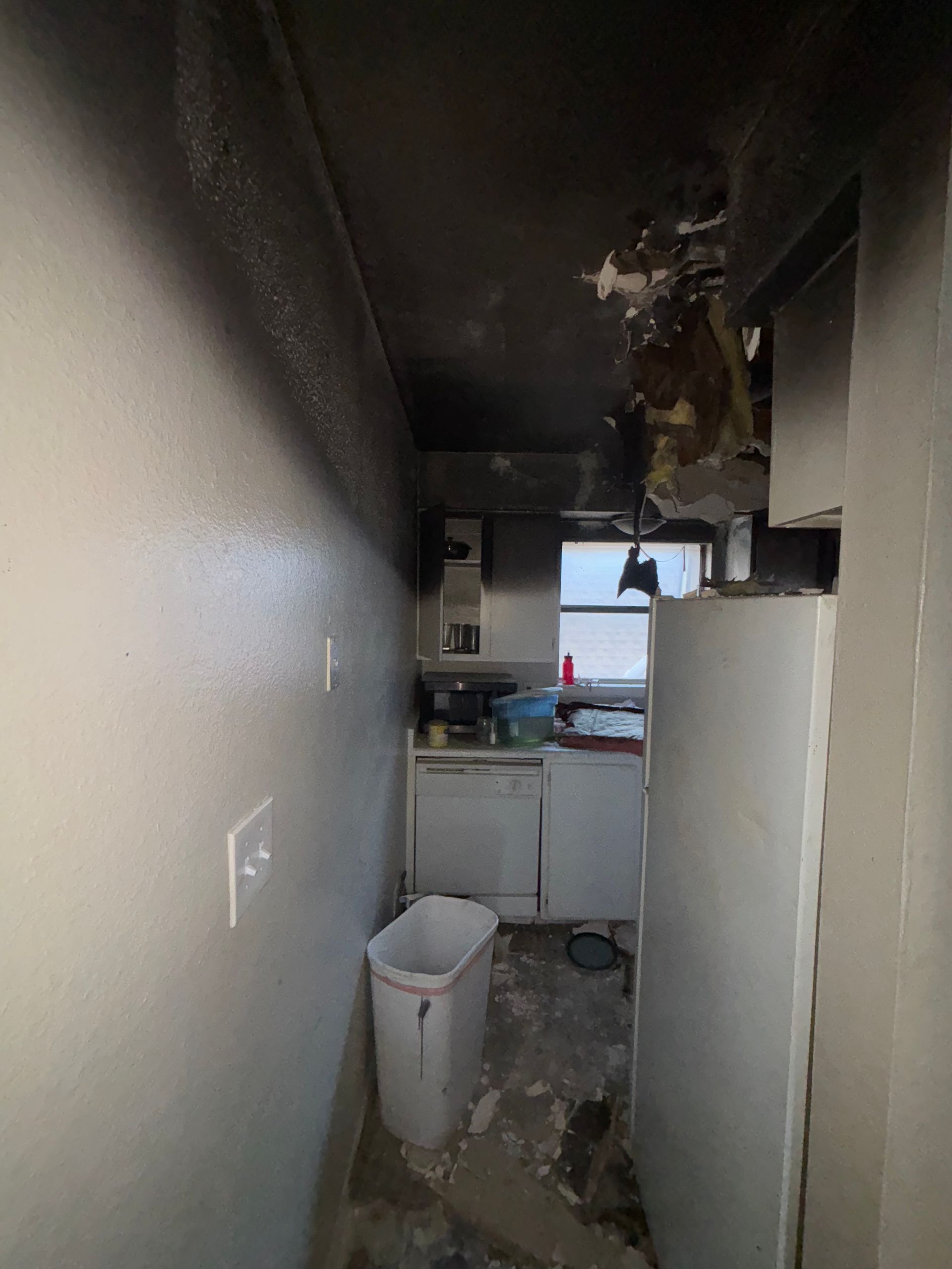 Narrow damaged kitchen hallway with debris, a trash bin, and a white refrigerator by a window.