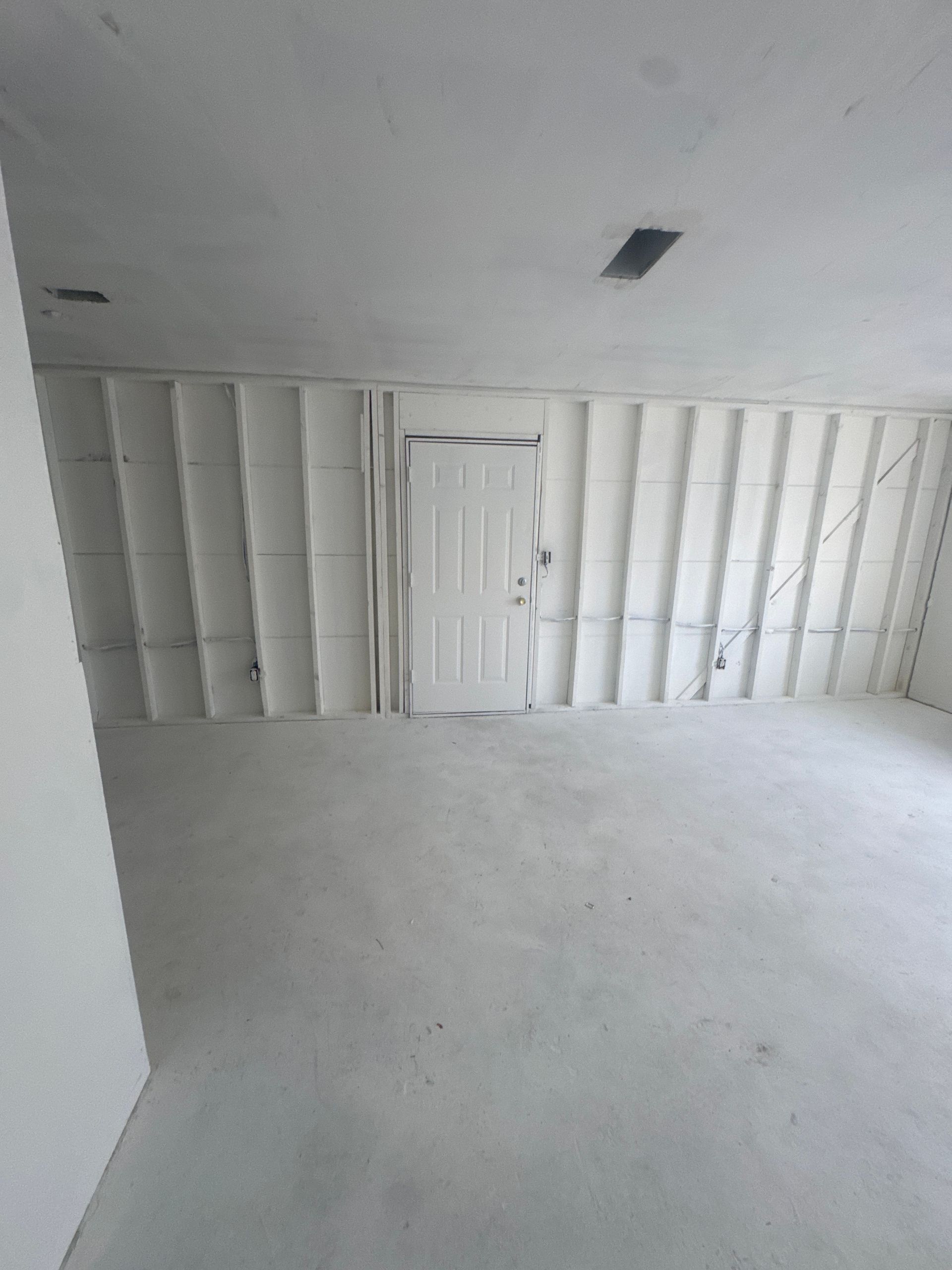 Empty unfinished room with white walls, exposed studs, and a closed white door on the far wall.