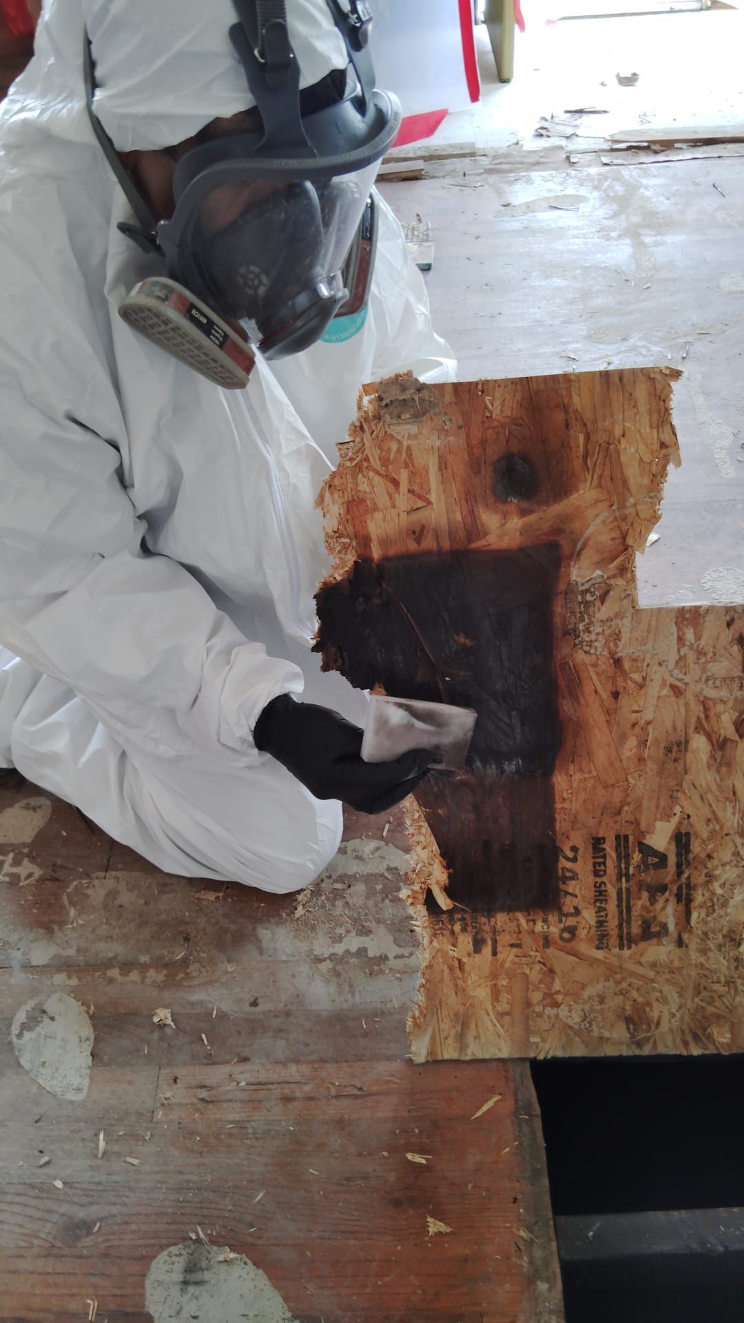 Worker in white protective gear examines a burnt floor section with a dark patch and plywood sheet nearby.
