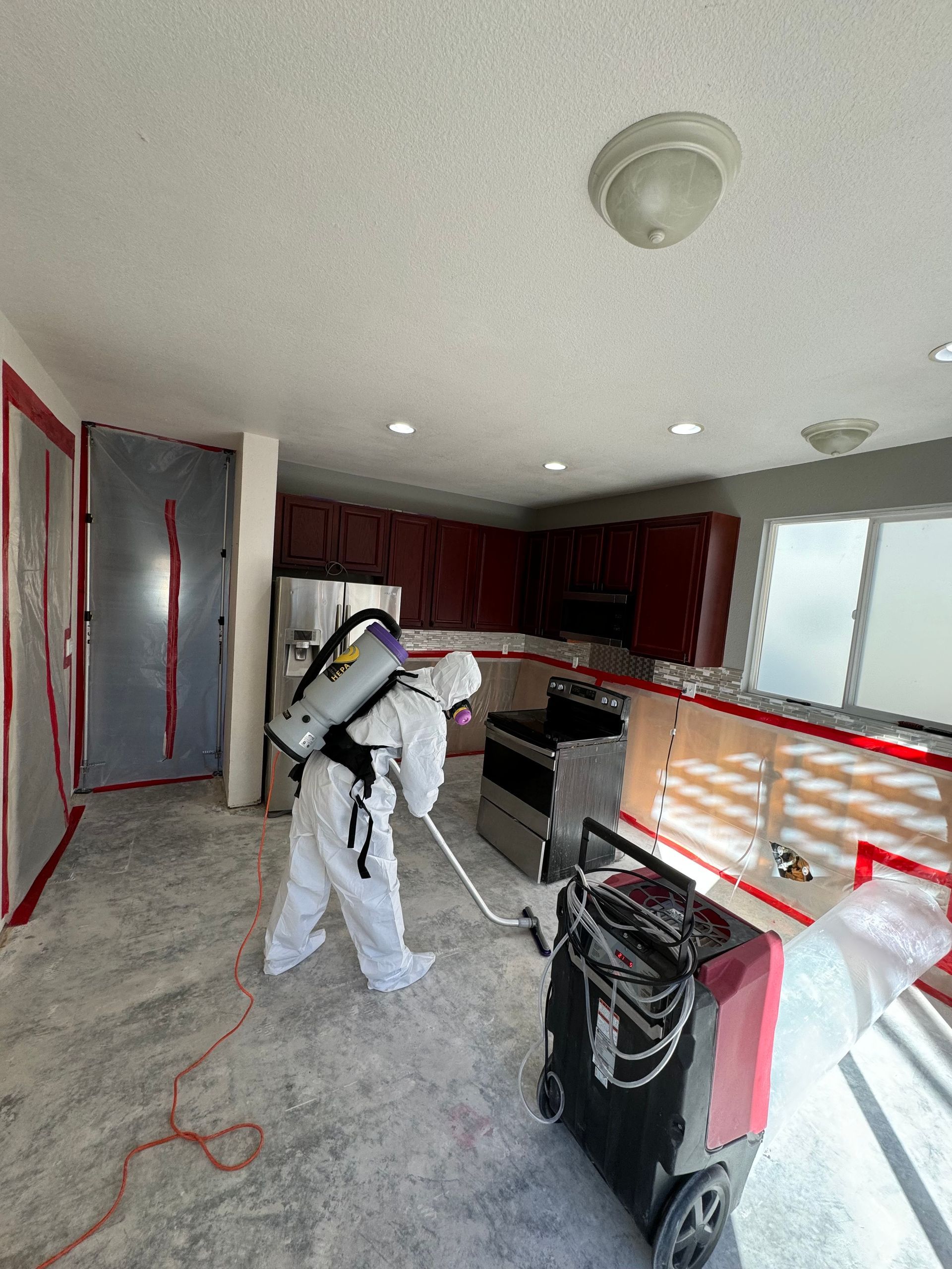 Worker sanding kitchen cabinets in a partially renovated room with tools, dust, and red cabinetry.
