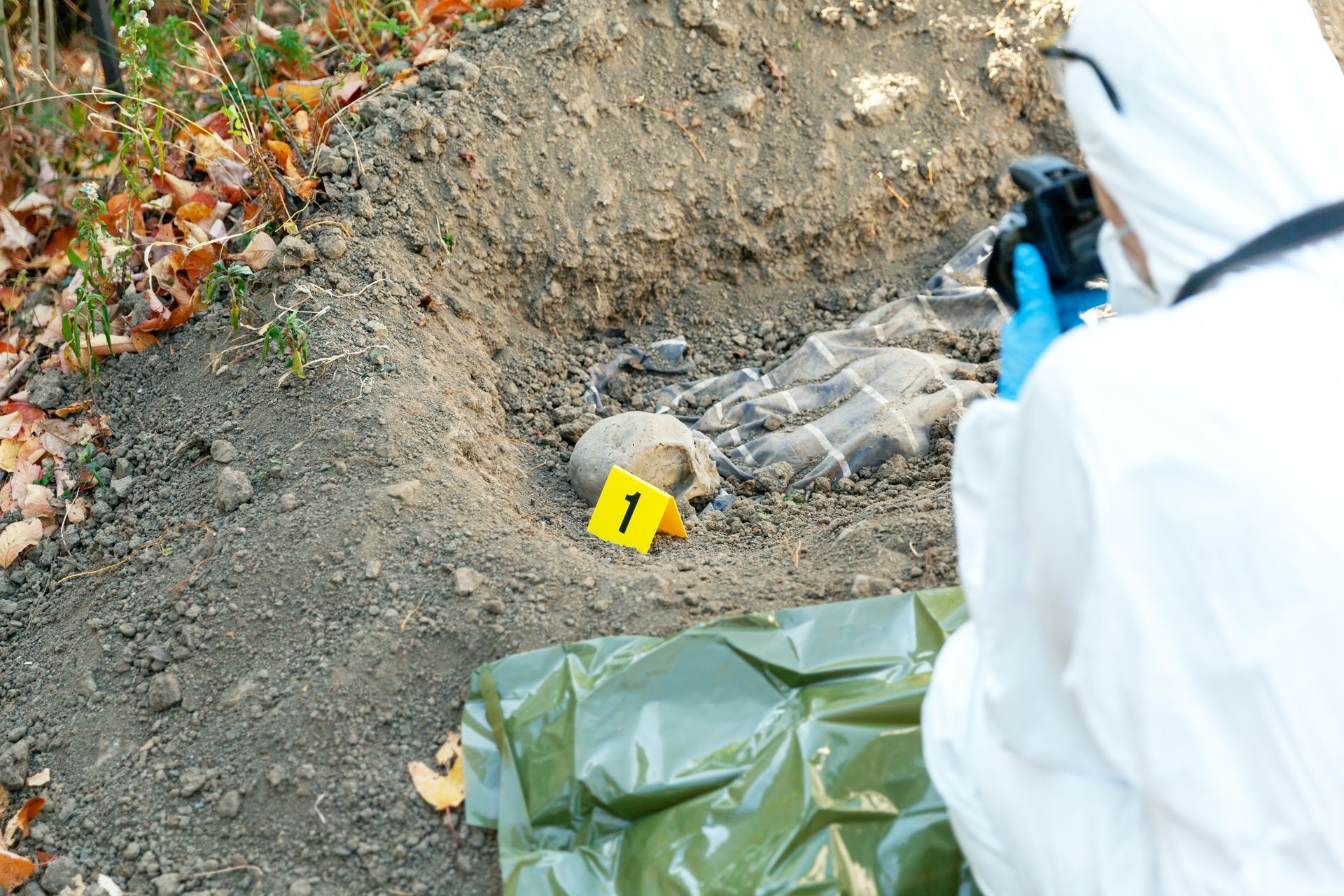 Person in white protective suit examining a marked area beside a wrapped object outdoors