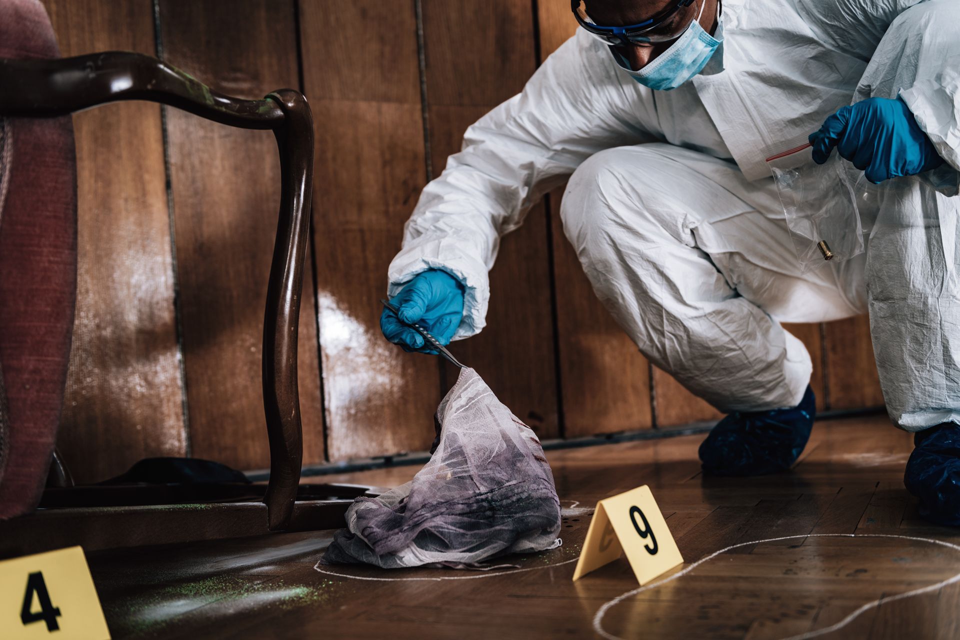 Crime scene investigator kneels beside evidence markers 4 and 9, examining a plastic-covered item on a wooden floor.