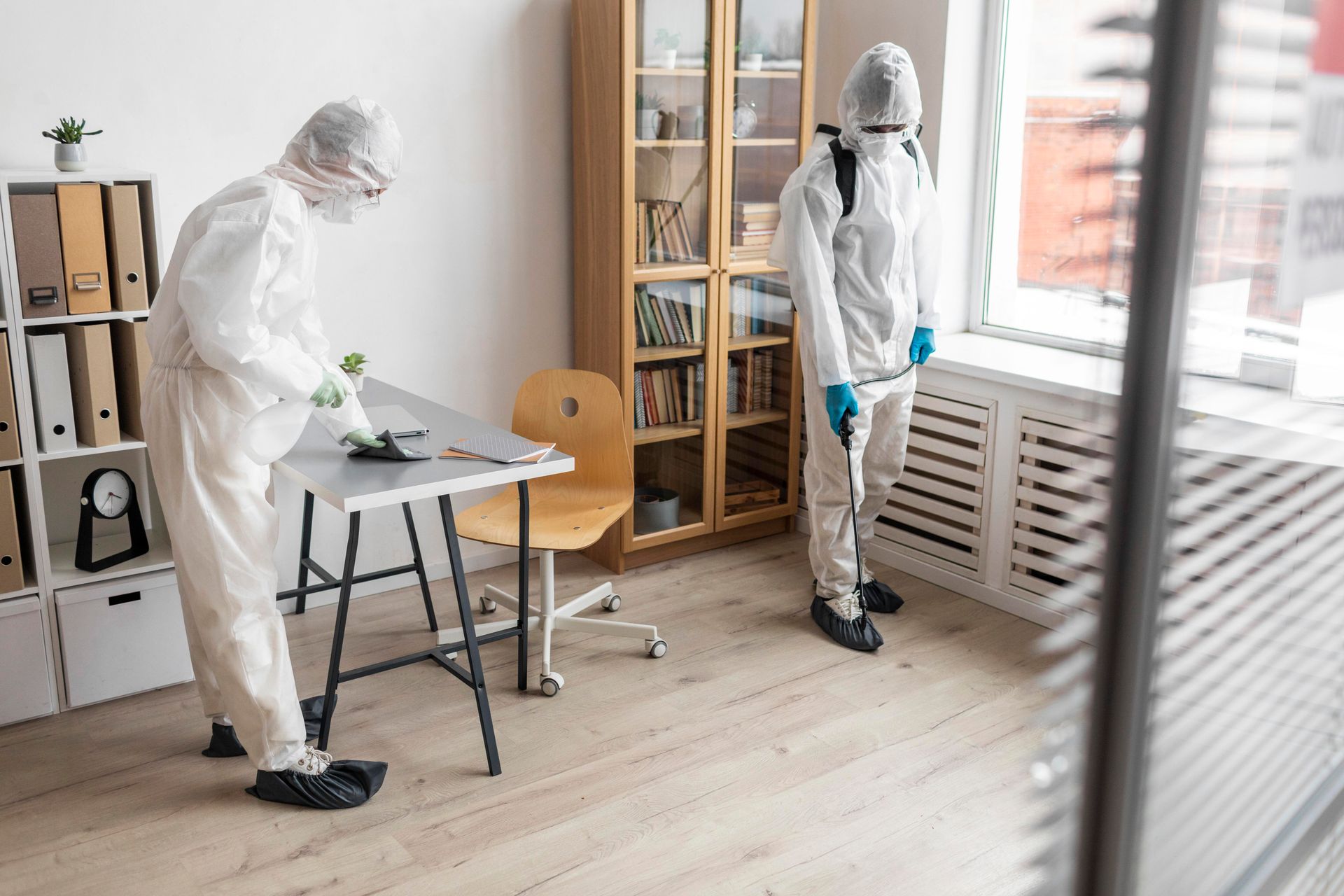 Two people in hazmat suits disinfecting a bright room near a window and table
