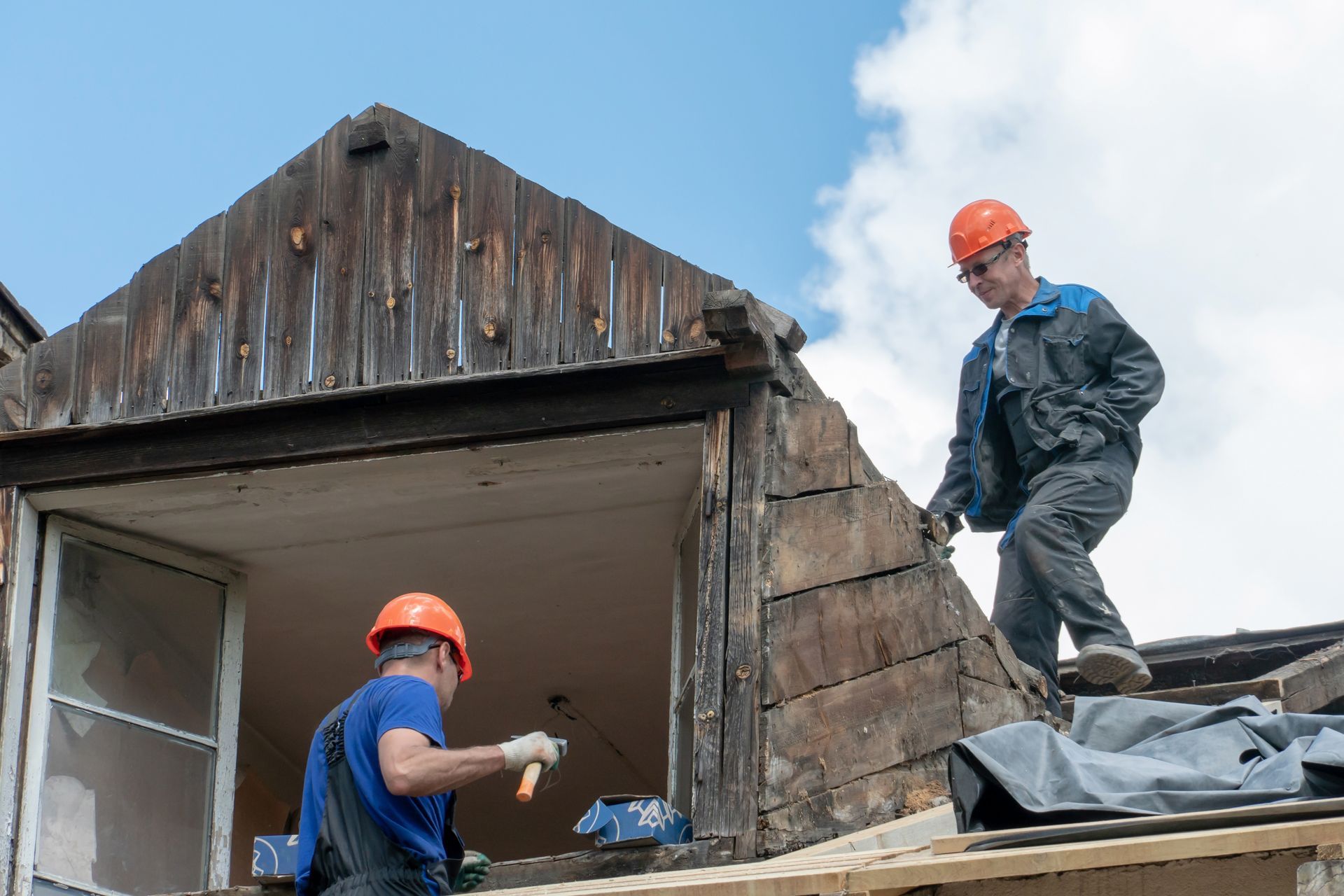 Two construction workers on a roof repairing a damaged building under a cloudy sky