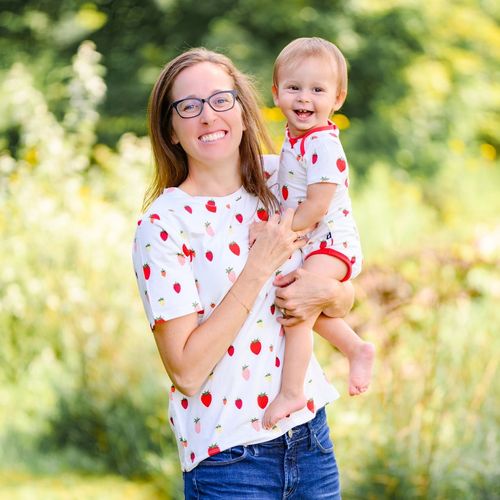 A woman and a young child in matching strawberry-print shirts smile while outdoors in a grassy, sunlit garden.