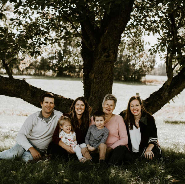 Six people pose for a portrait under a large tree in a grassy field on a sunny day.