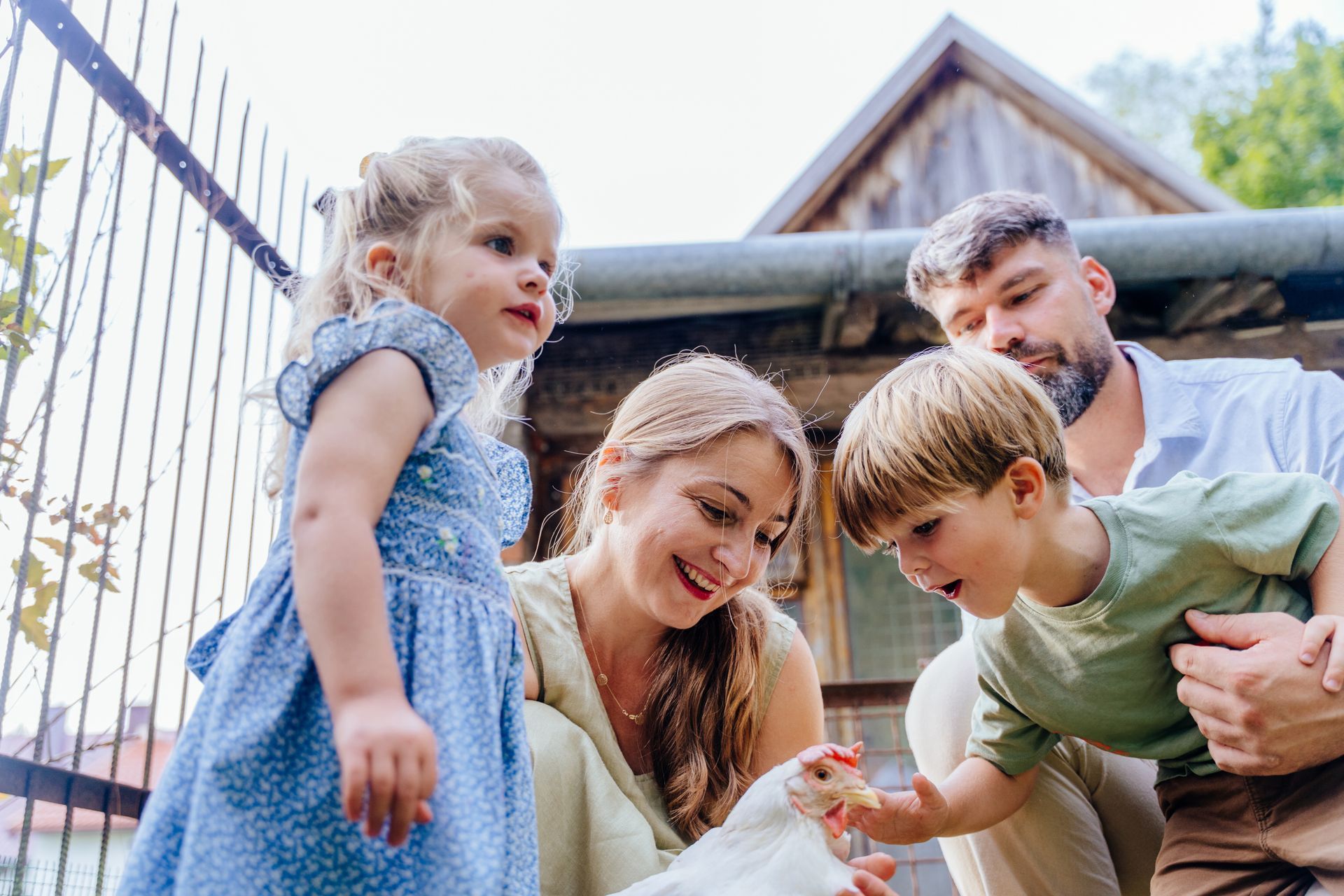 Adults and children crouch together outdoors, smiling and looking at something near the ground.
