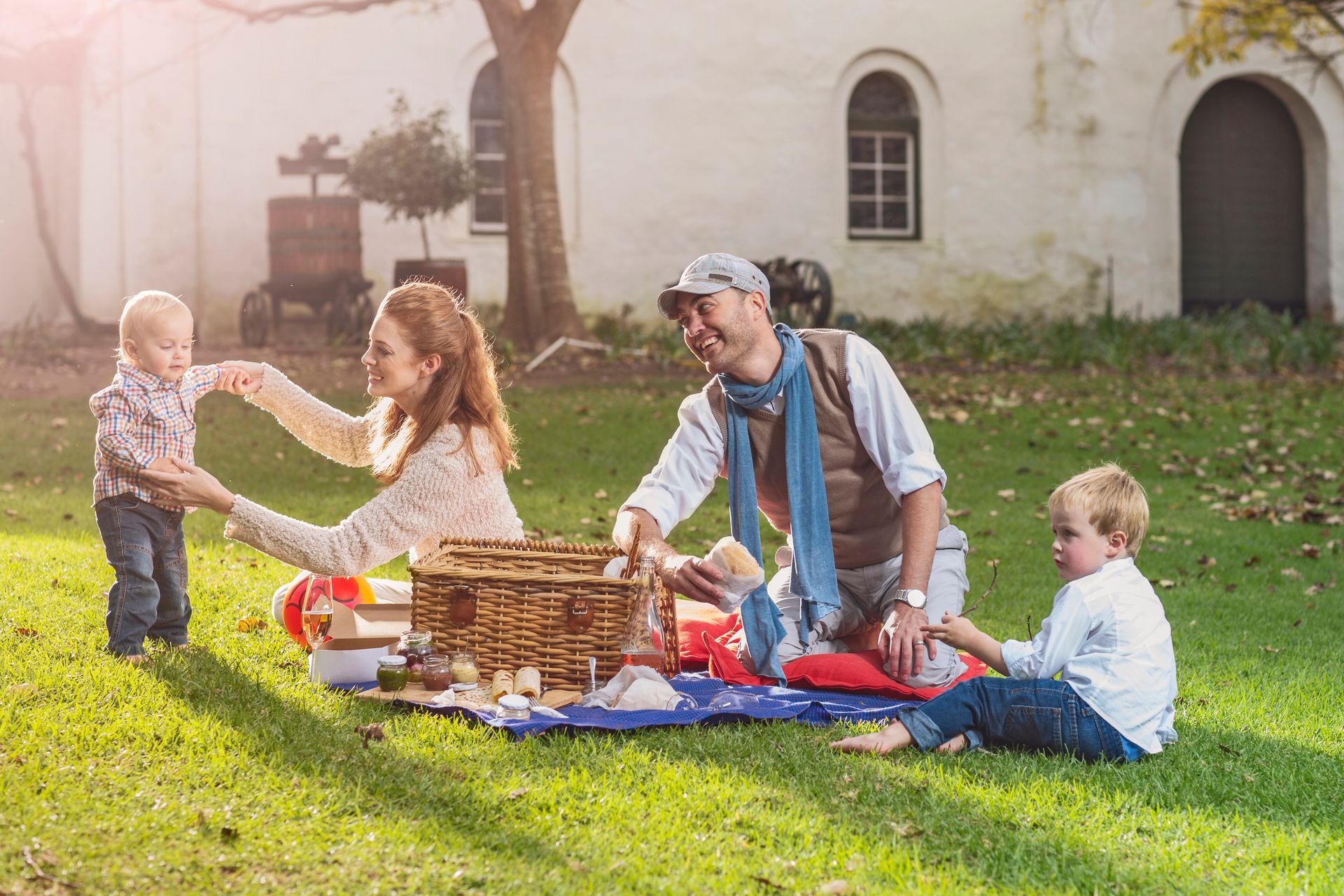 Family picnic on a sunny lawn, with adults and children sharing food on a blanket near a house.