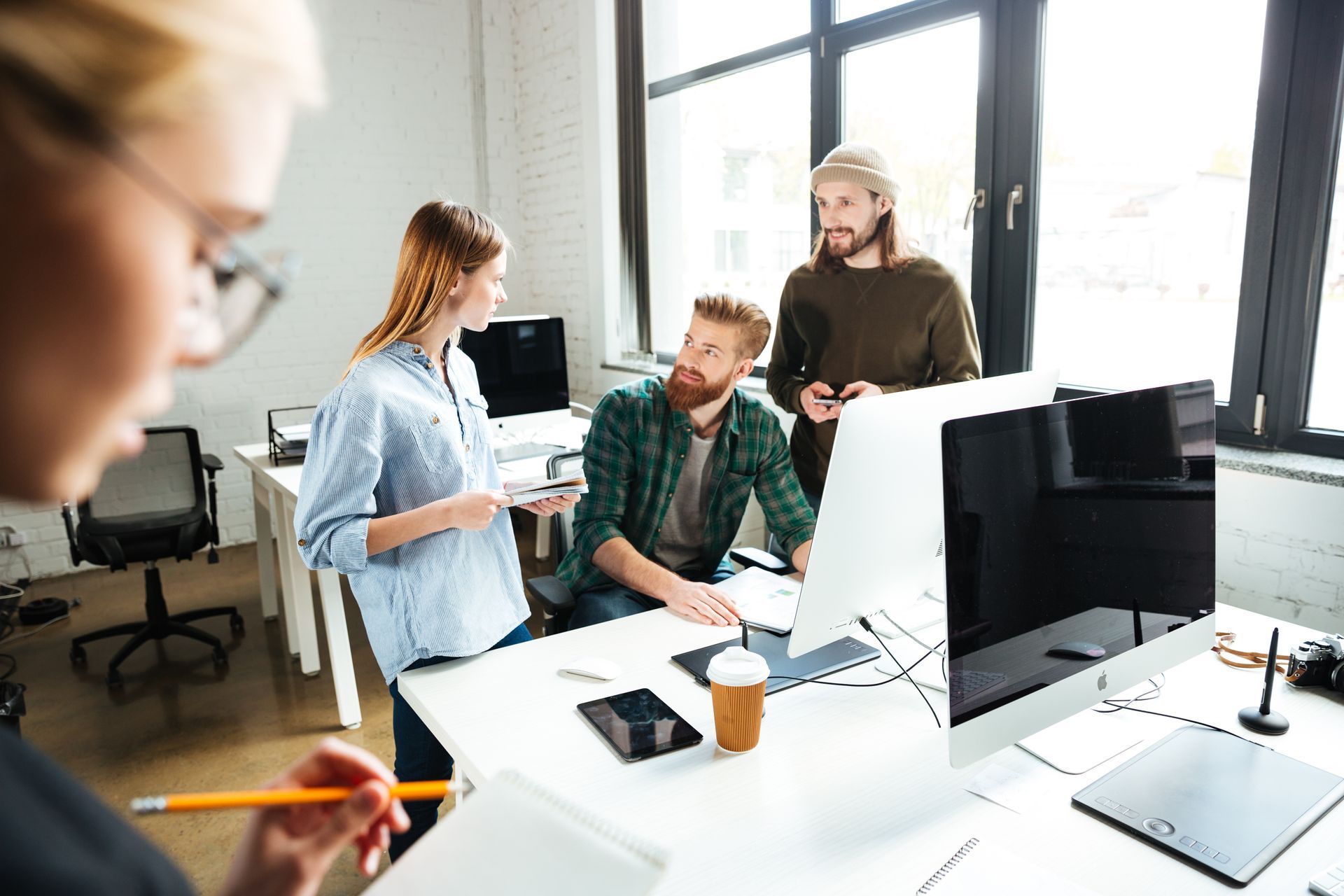 Colleagues discussing work around a bright office desk with computer monitors and notes