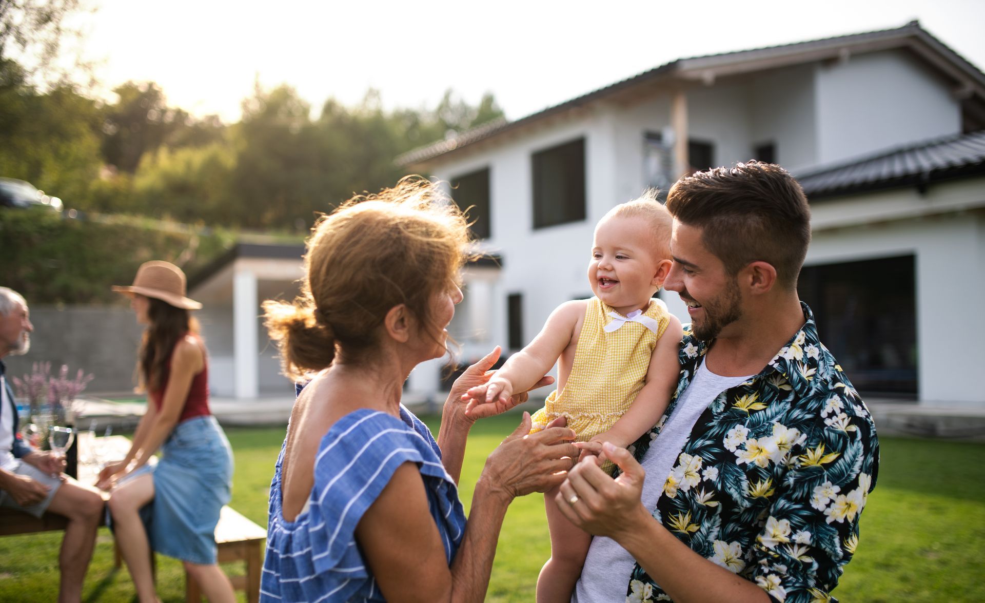 Family chatting outdoors in a sunny backyard, with a man holding a smiling child near a white house