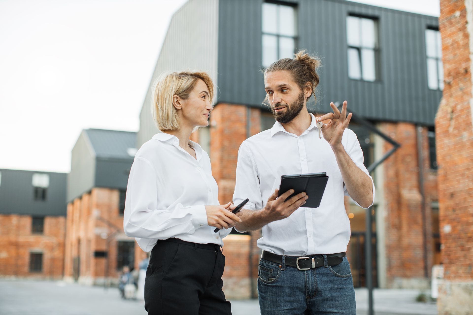 Two people in white shirts discuss a tablet outside modern brick buildings.