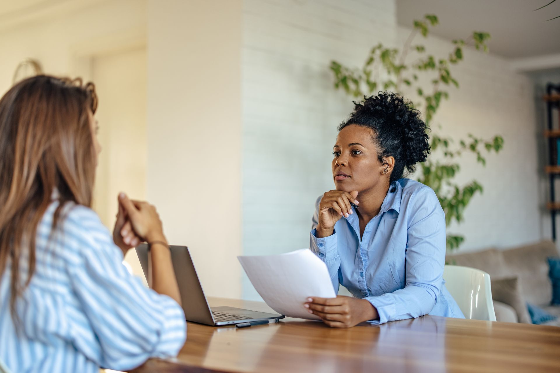 Two people in a meeting, one holding papers across a laptop on a wooden table