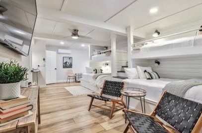 A modern bedroom with white bunk beds, wood floors, two woven lounge chairs, and a television on the wall.
