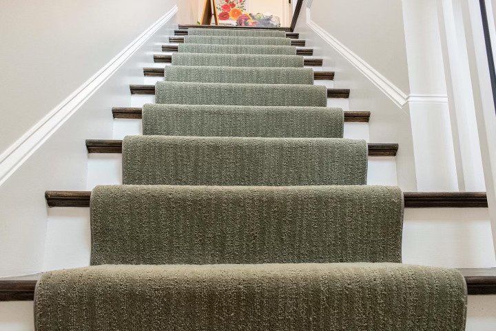 A straight staircase featuring dark wooden steps covered by a textured, olive-green carpet runner against white walls.
