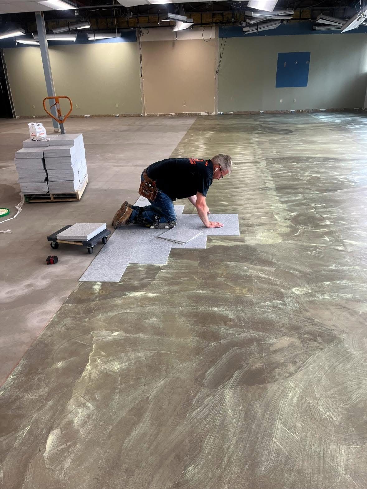 A person kneels on a concrete floor in a large, empty space, installing gray carpet tiles onto an adhesive-covered surface.