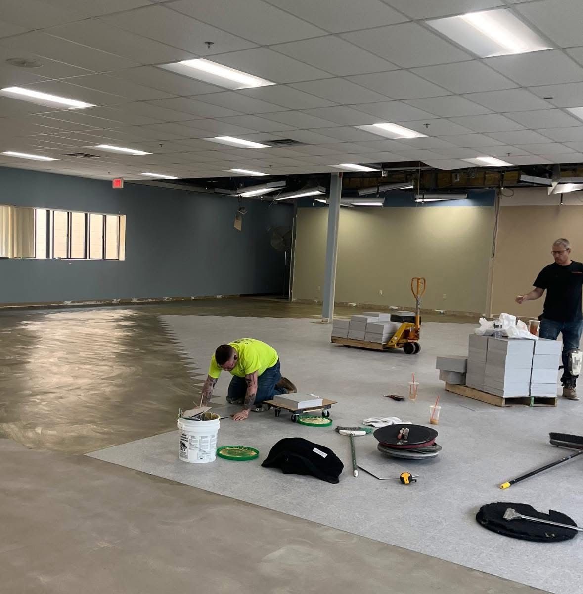 Two workers install gray flooring in a large, brightly lit commercial space with light-colored walls and ceiling tiles.