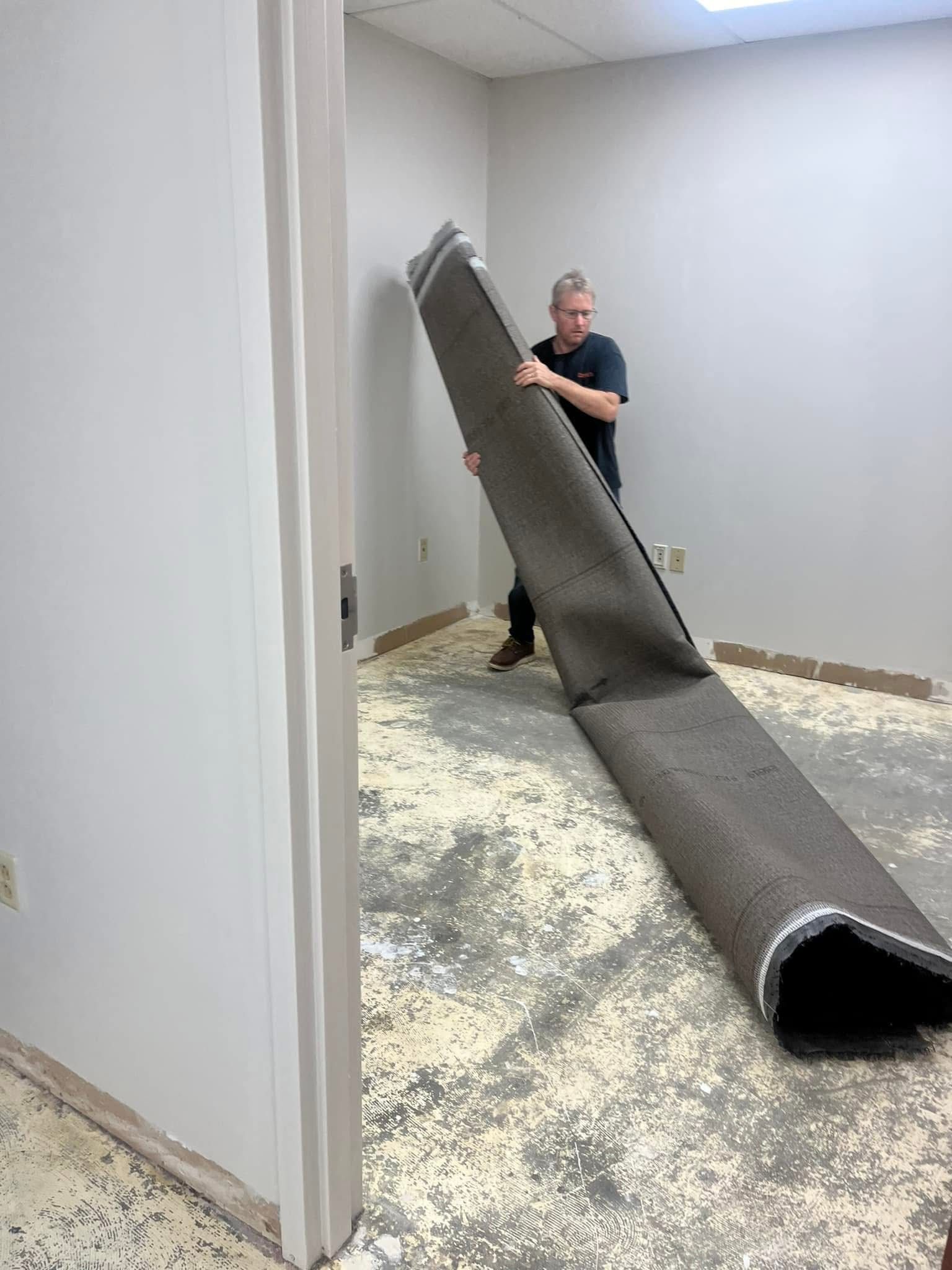 A person lifts a long roll of gray carpet in an office room with bare, debris-covered flooring.