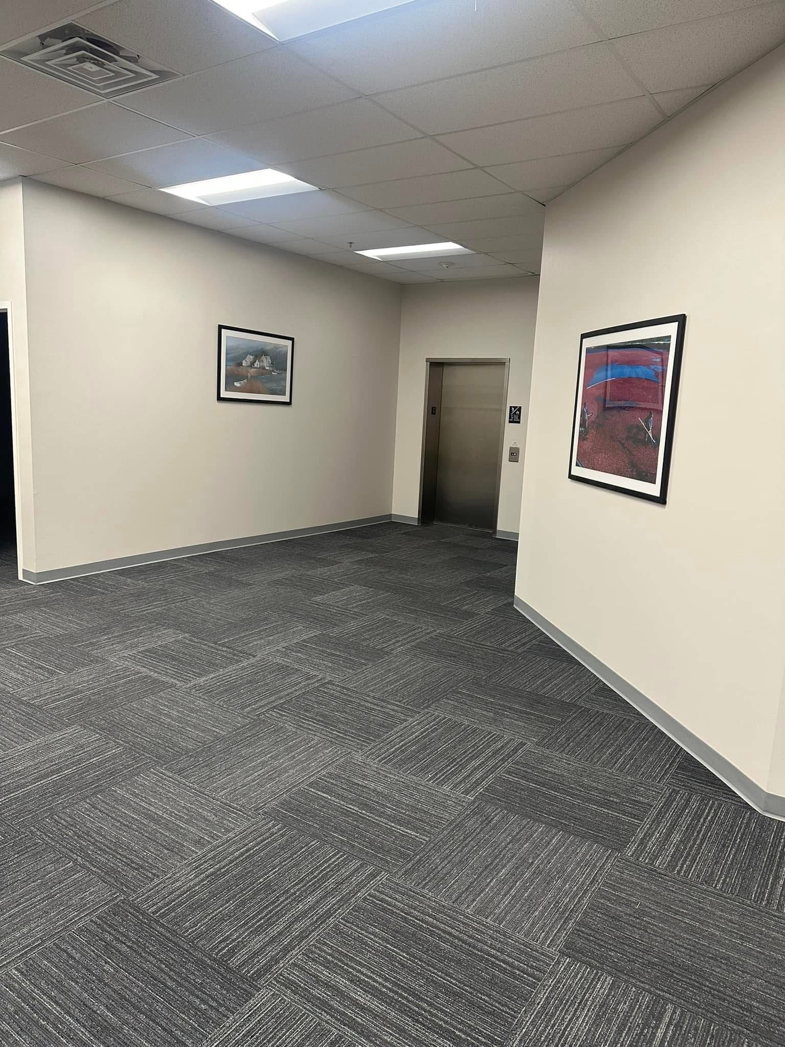A hallway with gray carpet, beige walls, two framed prints, and an elevator door at the end.