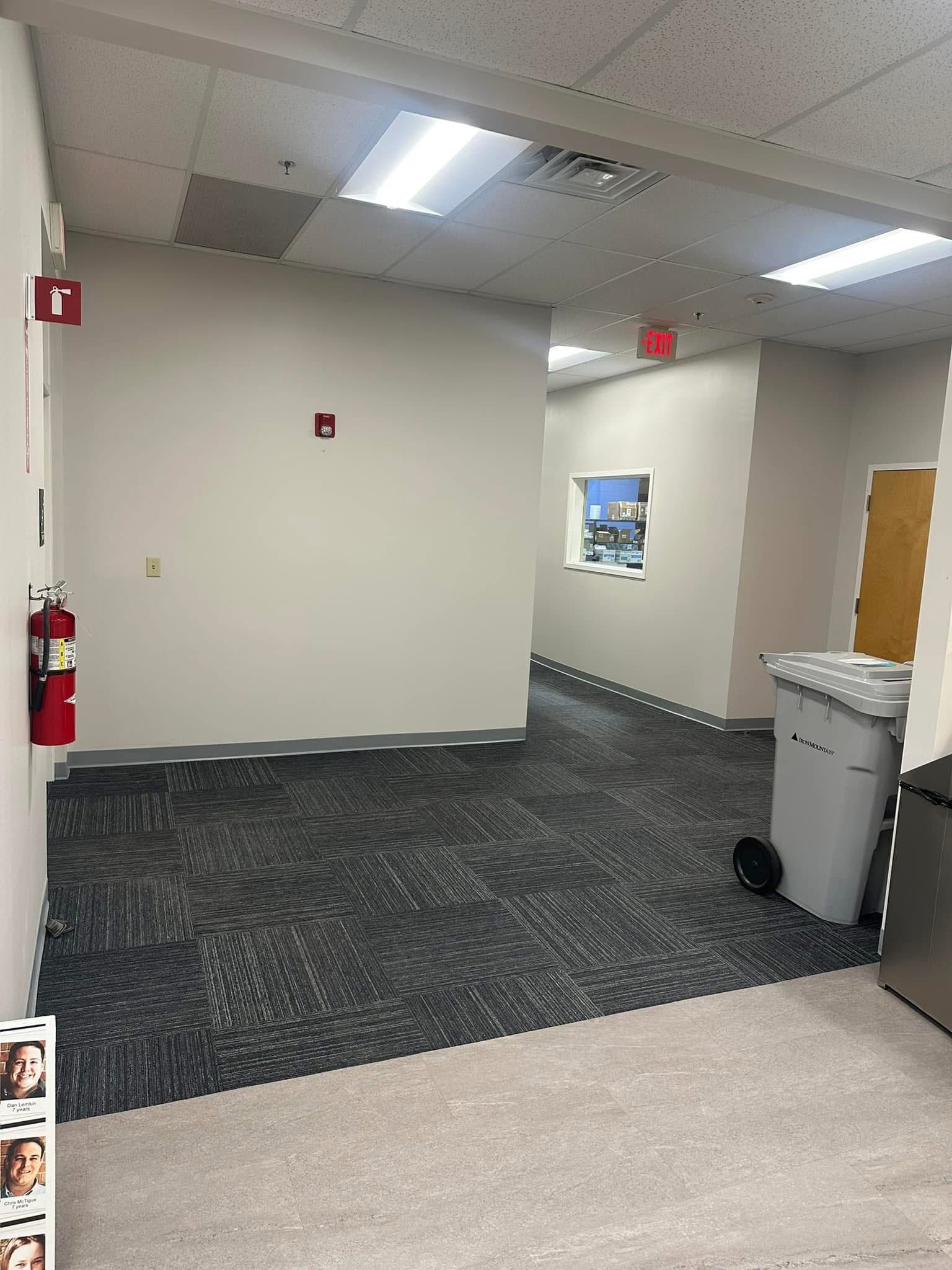 An interior office hallway with carpeted flooring, a fire extinguisher on the wall, and a trash bin in the corner.