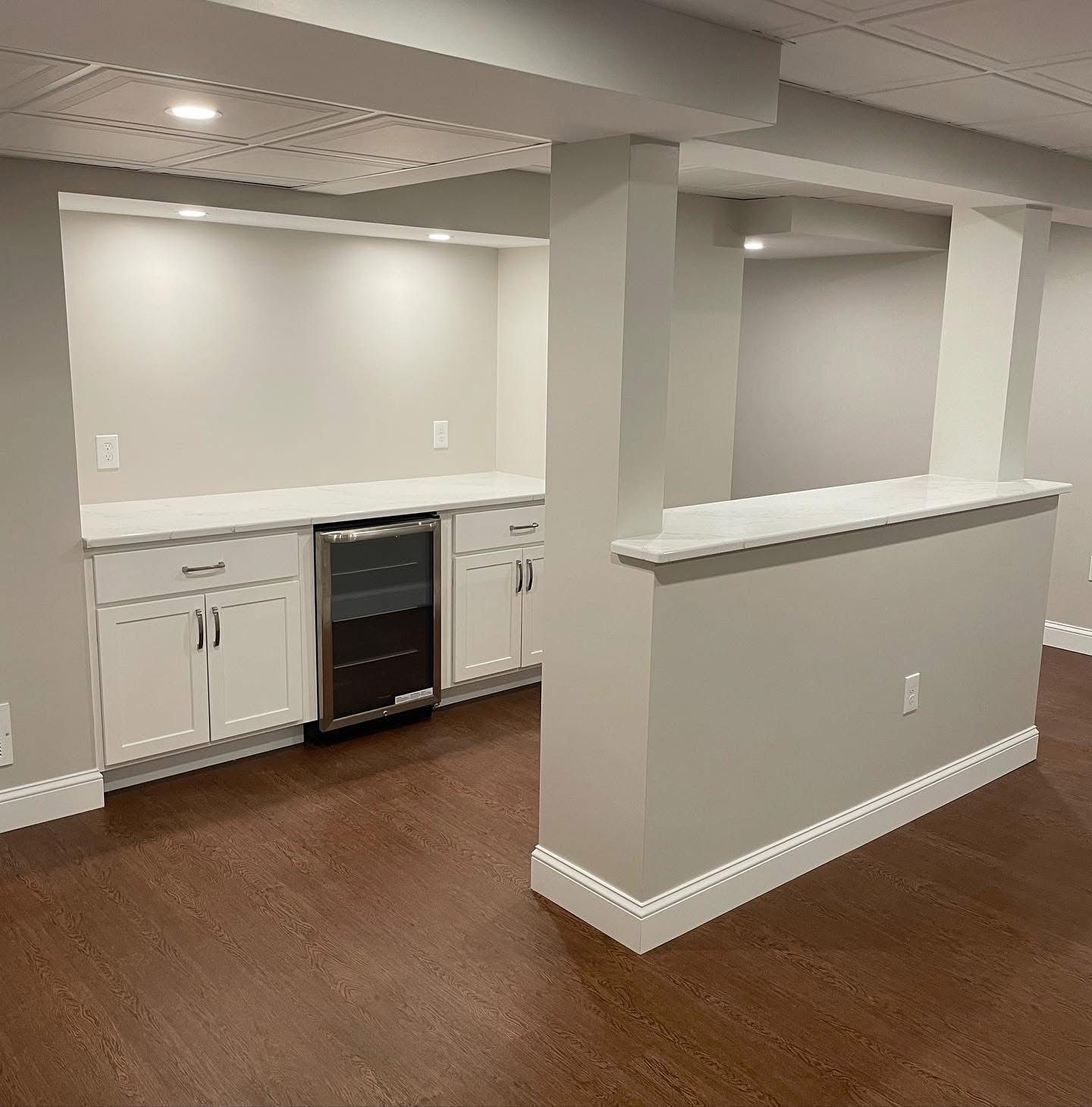A home basement area featuring white cabinets, a small wine cooler, and a counter divider against a neutral wall.