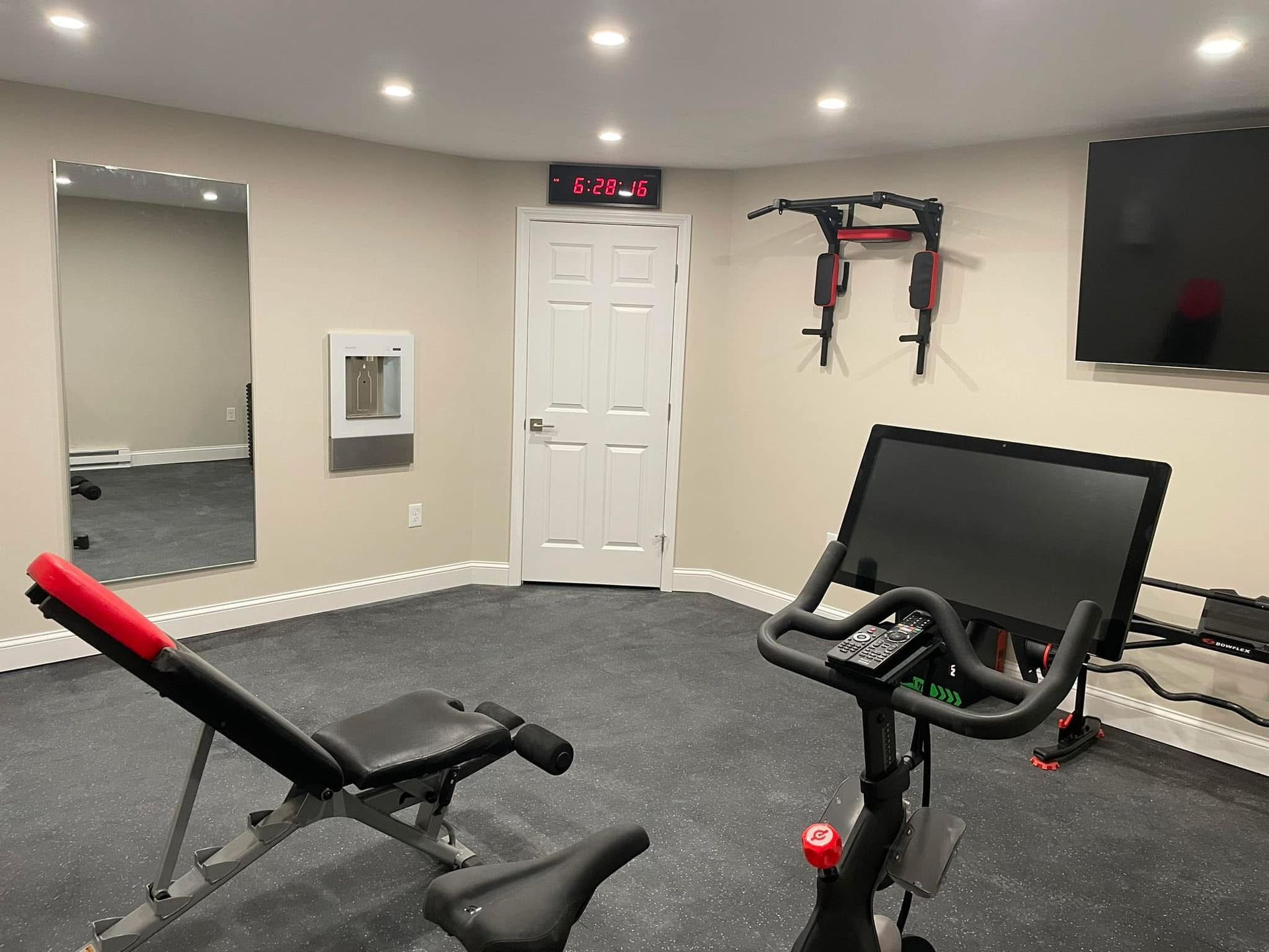 A home gym with a stationary bike, weight bench, pull-up bar, and wall mirror in a room with light walls and dark flooring.