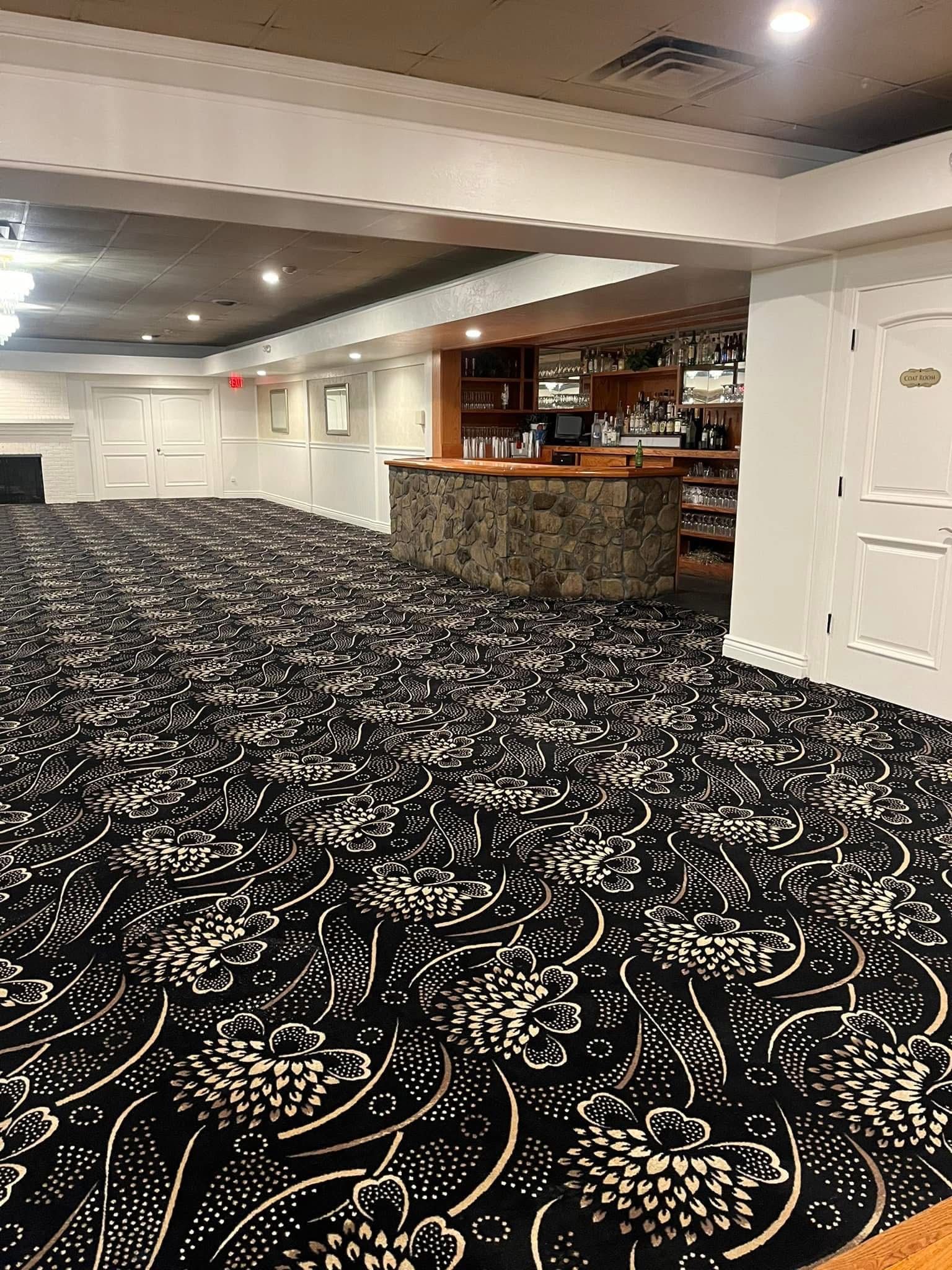 A spacious room with a patterned black and gold carpet, a stone bar, and white paneled walls under recessed lighting.