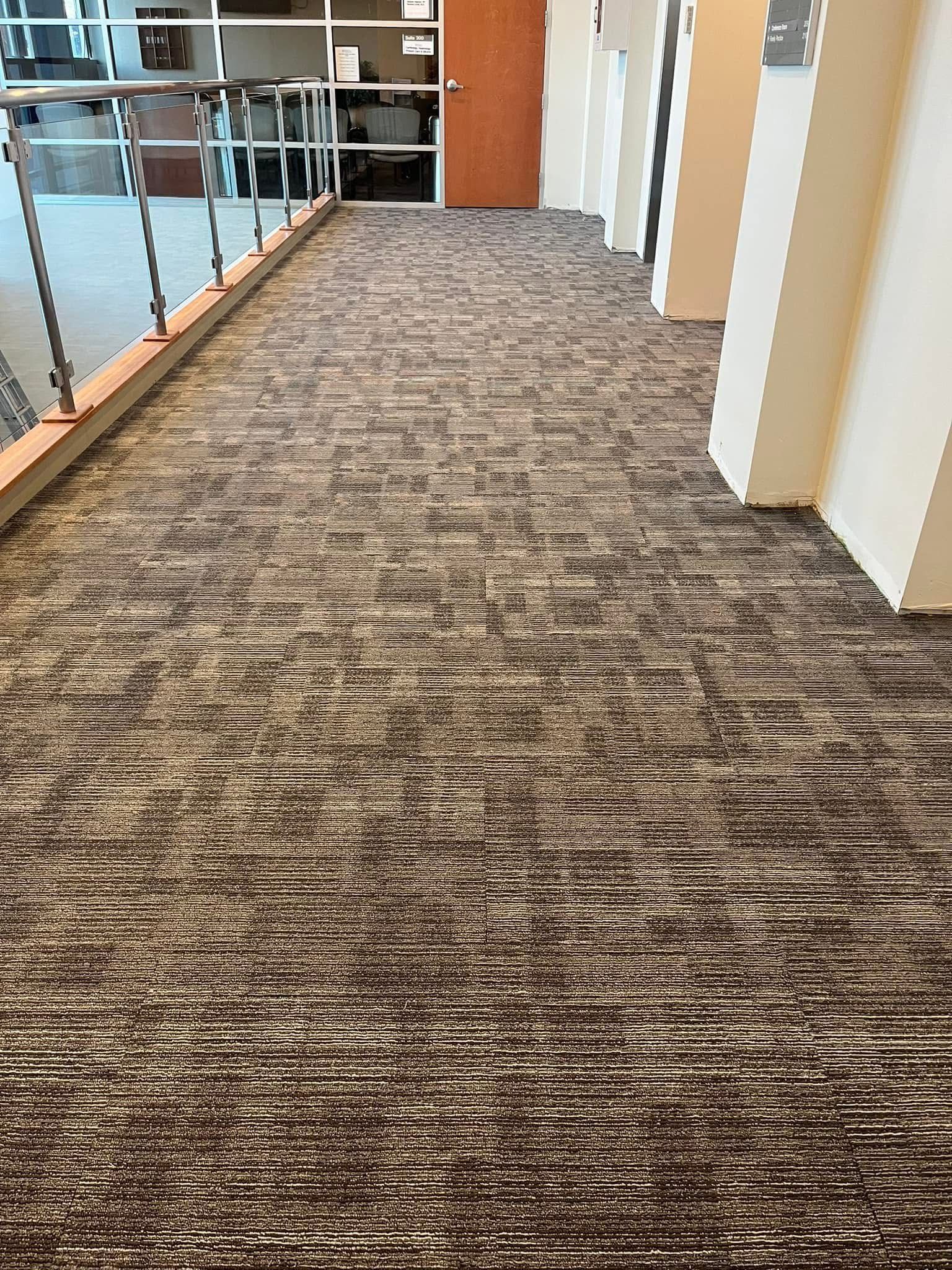 A hallway with textured, grey-and-brown patterned carpet, a glass railing on the left, and white pillars on the right.