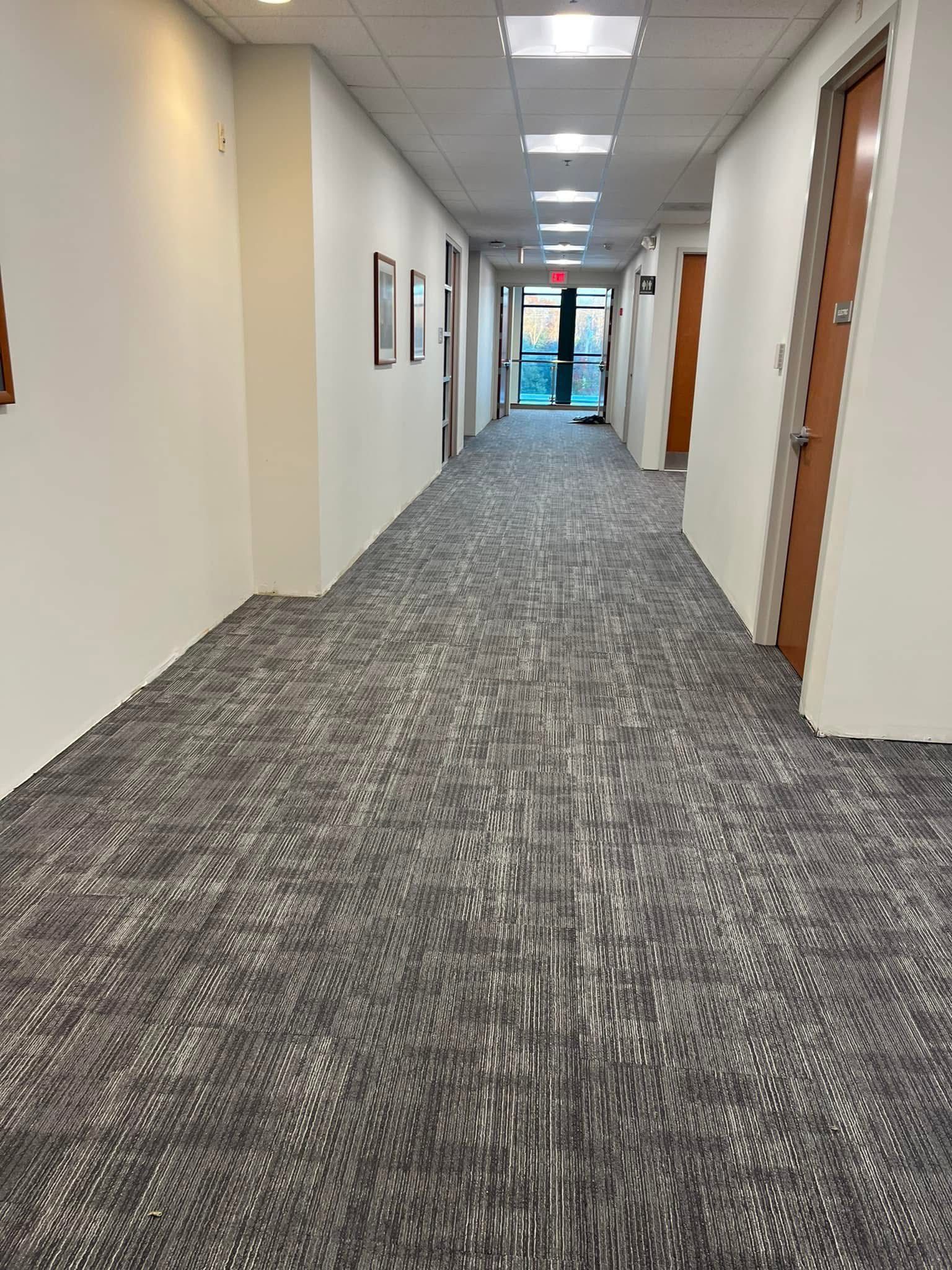 A narrow office hallway with white walls, a gray patterned carpet, drop ceiling lights, and several closed wooden doors.