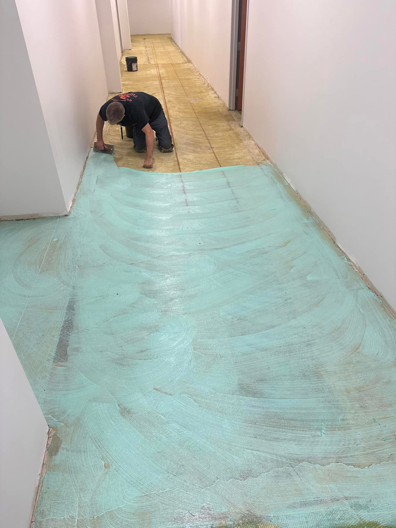 A worker kneels on a long hallway floor, applying a light blue adhesive to the subfloor for new flooring installation.