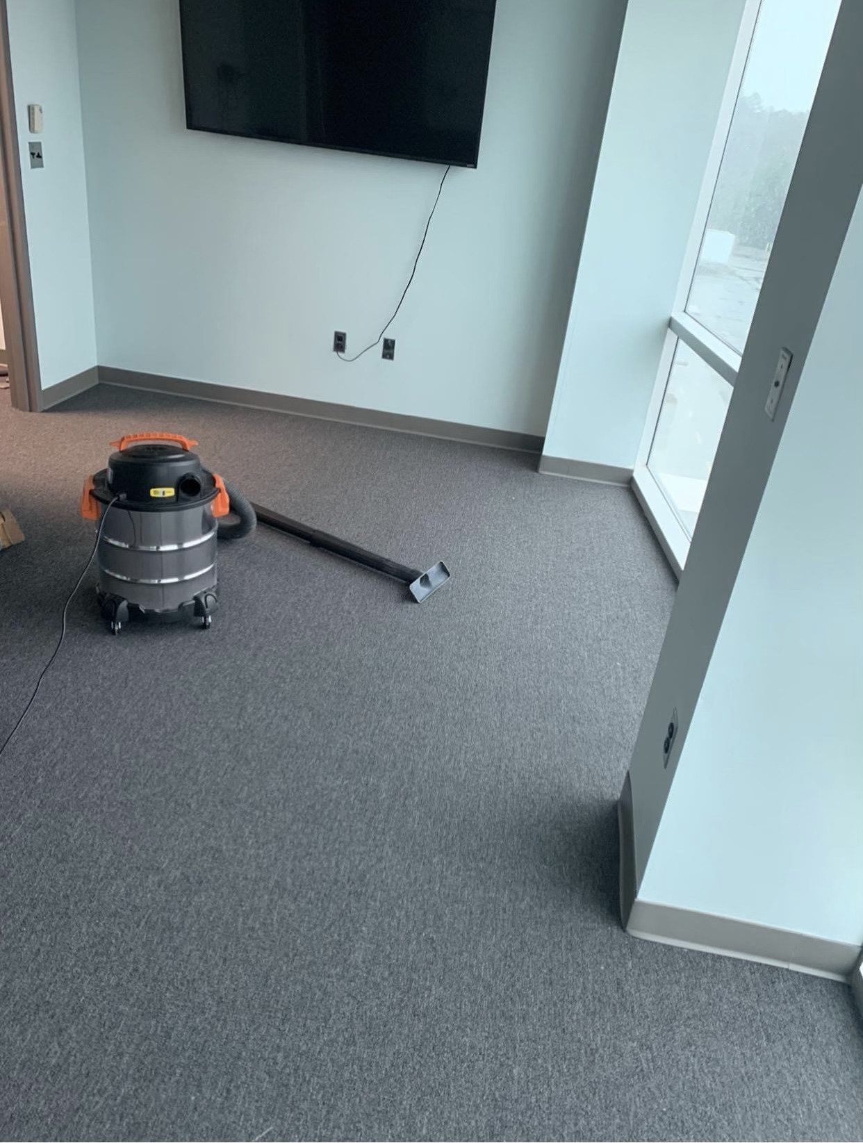 A shop vacuum with its wand resting on a gray office carpet near a wall-mounted TV and large windows.