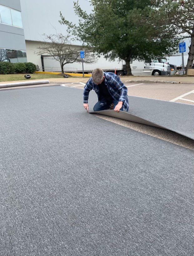 A person in a blue plaid shirt kneels in an outdoor parking lot, laying down a large roll of grey textured flooring material.