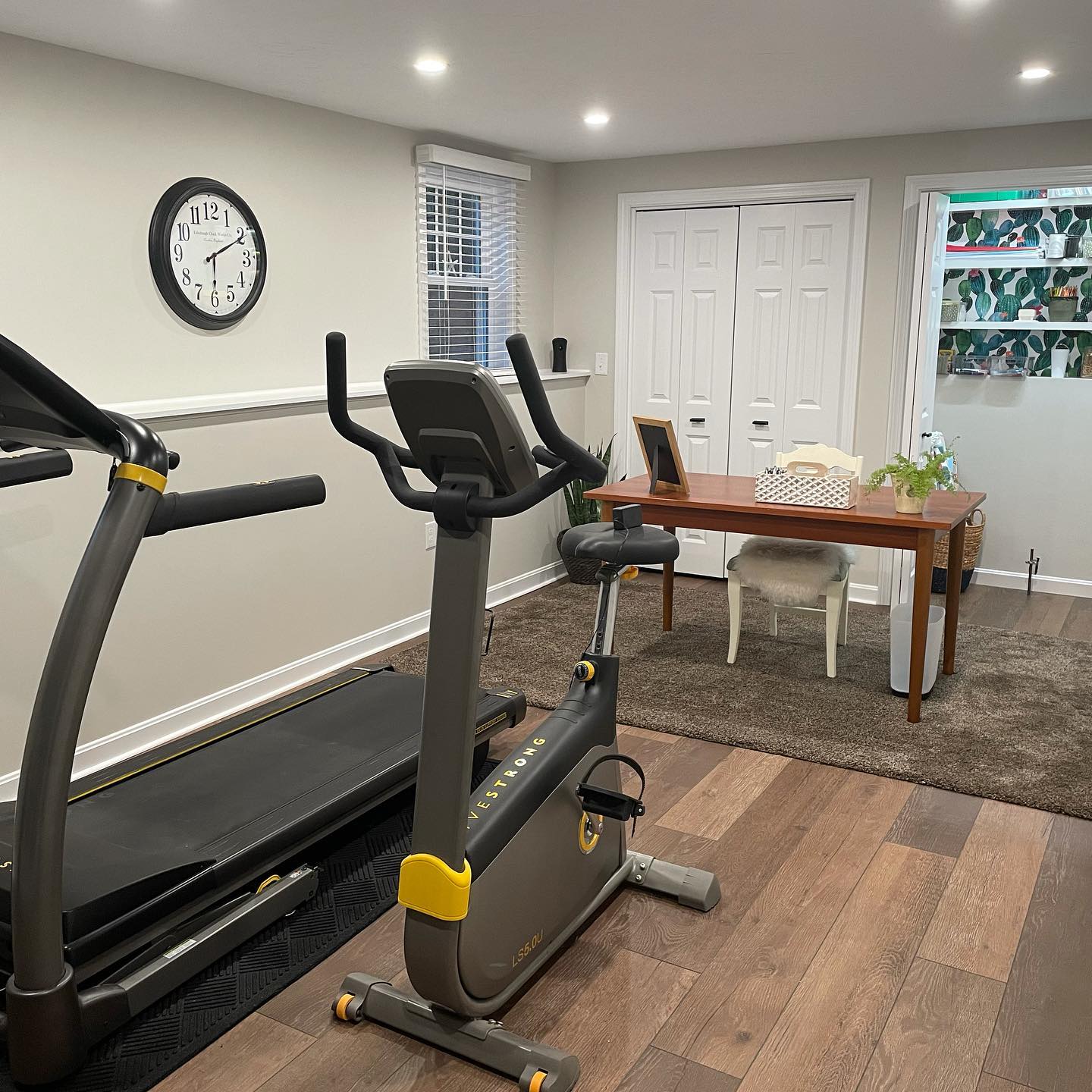 A treadmill and stationary bike in a room with light gray walls, a wooden desk with a chair, and a clock on the wall.