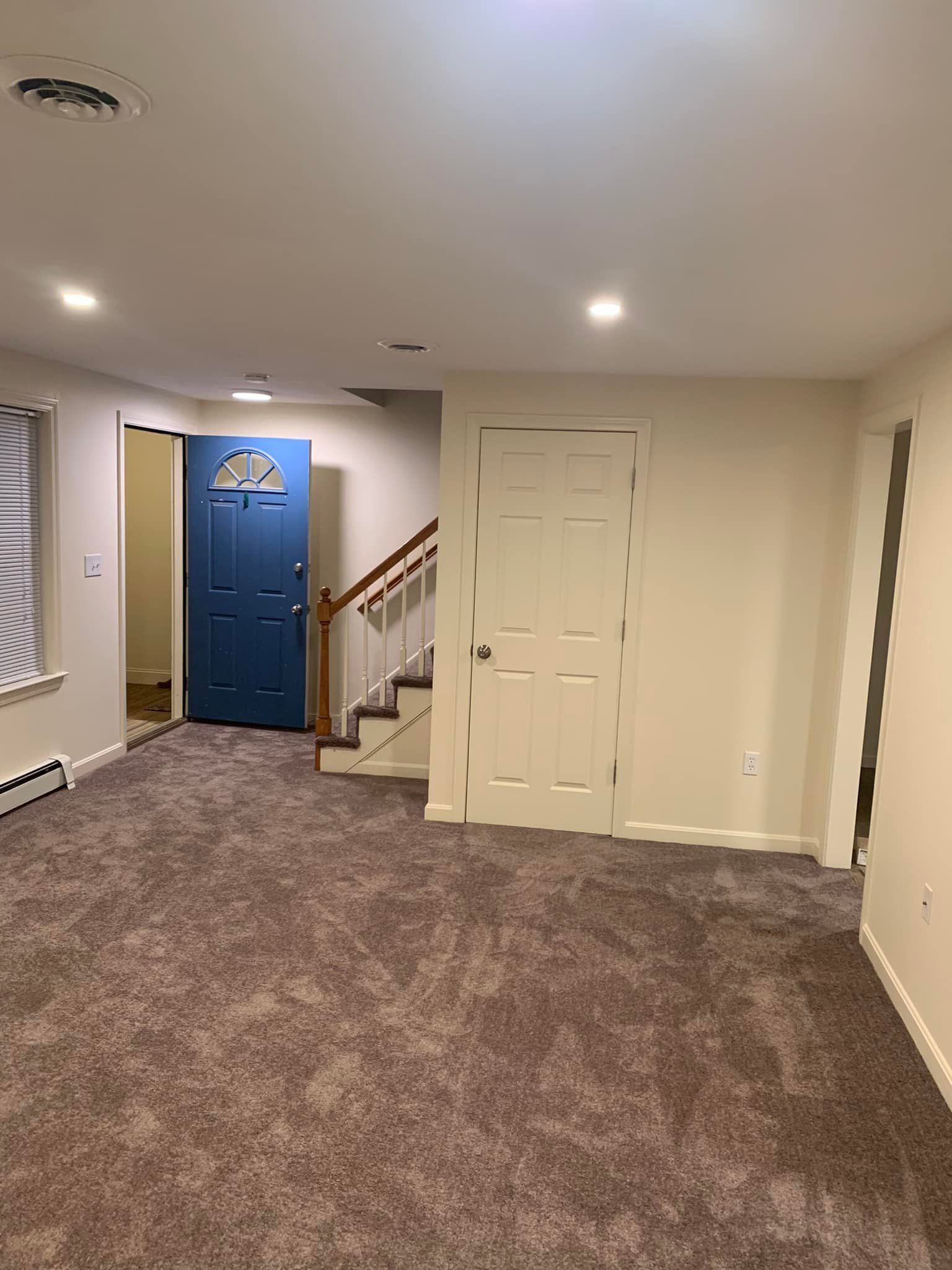 An interior view of a carpeted room featuring a blue front door, an adjacent staircase, and a white closed door.