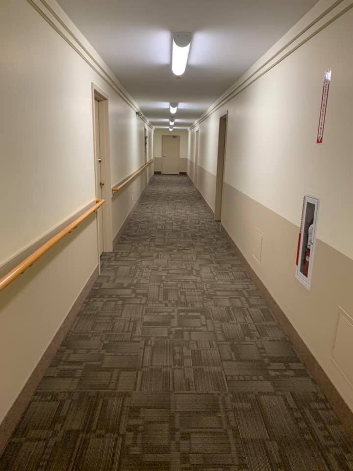 A long, dimly lit indoor hallway with beige walls, patterned carpet, and wooden handrails on the sides.