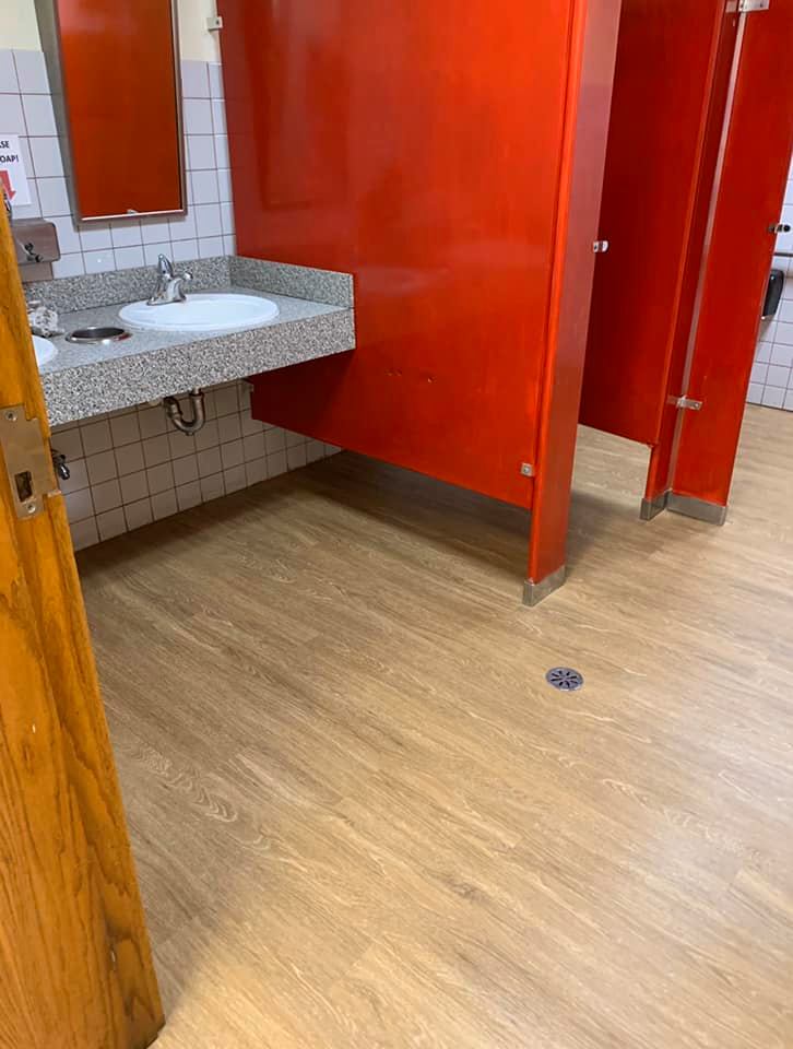 A public restroom with light wood-look flooring, a gray granite-style sink countertop, and bright orange stall doors.