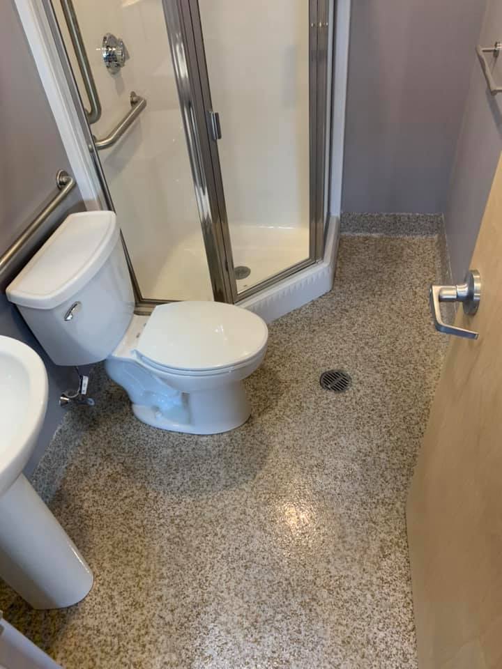 An accessible bathroom features a white toilet, corner shower with grab bars, pedestal sink, and a floor drain.