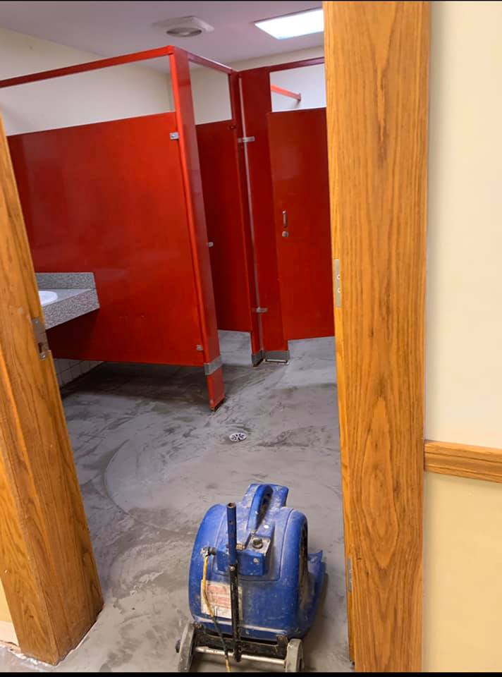 A blue industrial floor dryer sits in the doorway of a public restroom with red stall partitions and exposed concrete floor.
