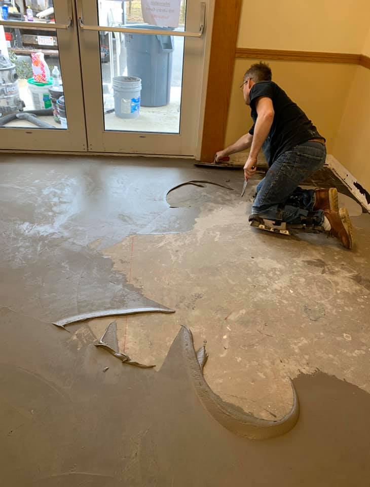 A person kneeling on a floor, smoothing a layer of gray wet concrete over an unfinished subfloor near glass doors.