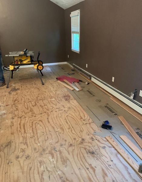 A room undergoing floor renovations with a table saw, exposed plywood subfloor, and pieces of new flooring.