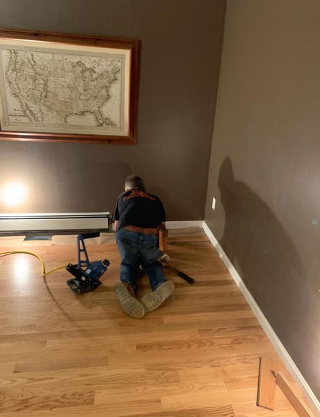 A professional installs hardwood flooring in a room with brown walls and a framed map on the wall.