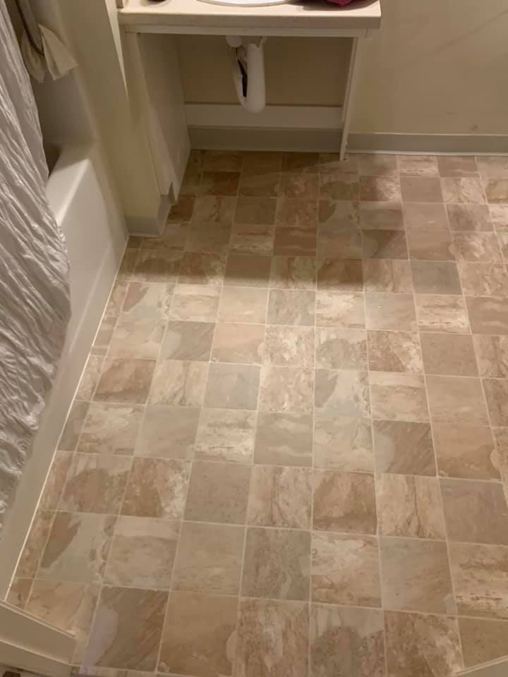 A high-angle shot of a beige, textured square tile bathroom floor next to a white bathtub and vanity.