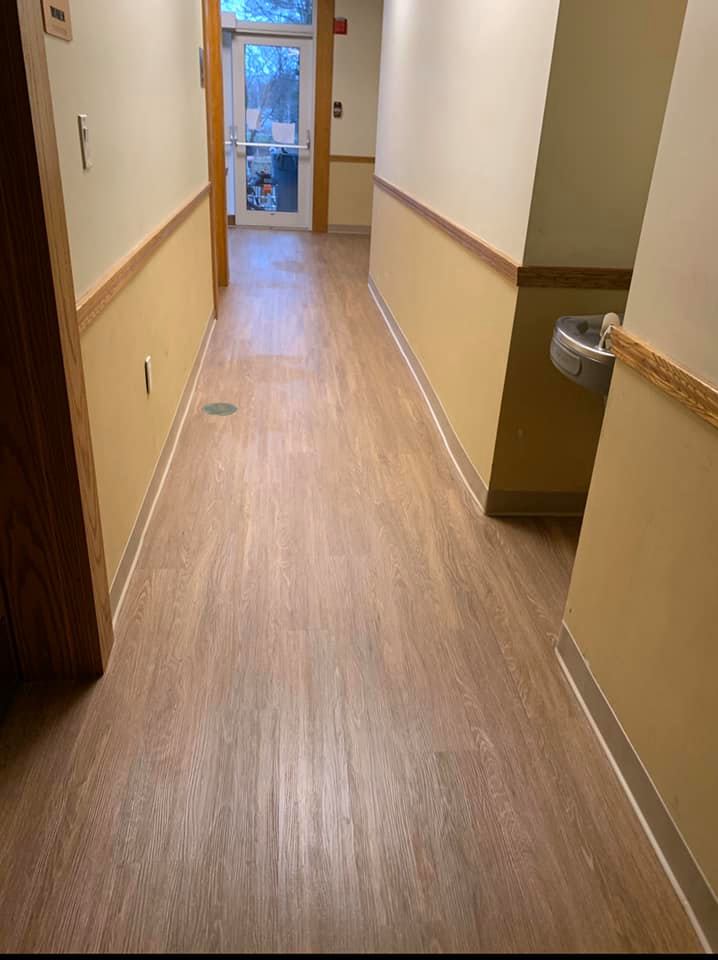 A hallway with light wood-look floors, tan walls with wooden trim, and a water fountain, leading to an exterior glass door.