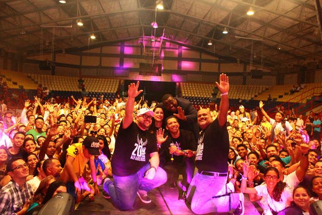 Un grupo de personas con camisetas negras idénticas, arrodilladas en un escenario, saludan a una gran multitud qu